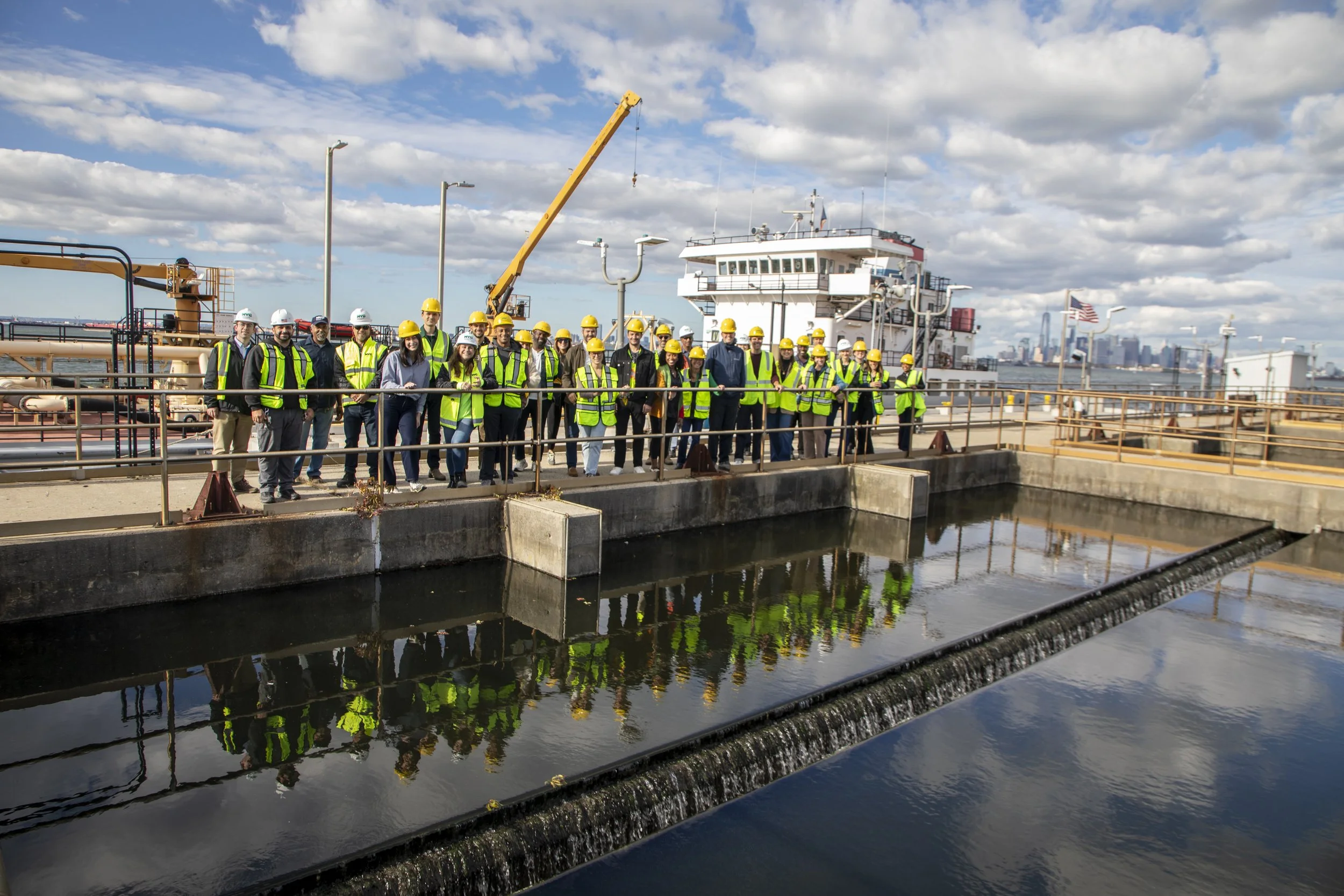Group of people wearing yellow and white safety helmets and reflective vests standing on a rooftop water treatment area with city skyline in the background.