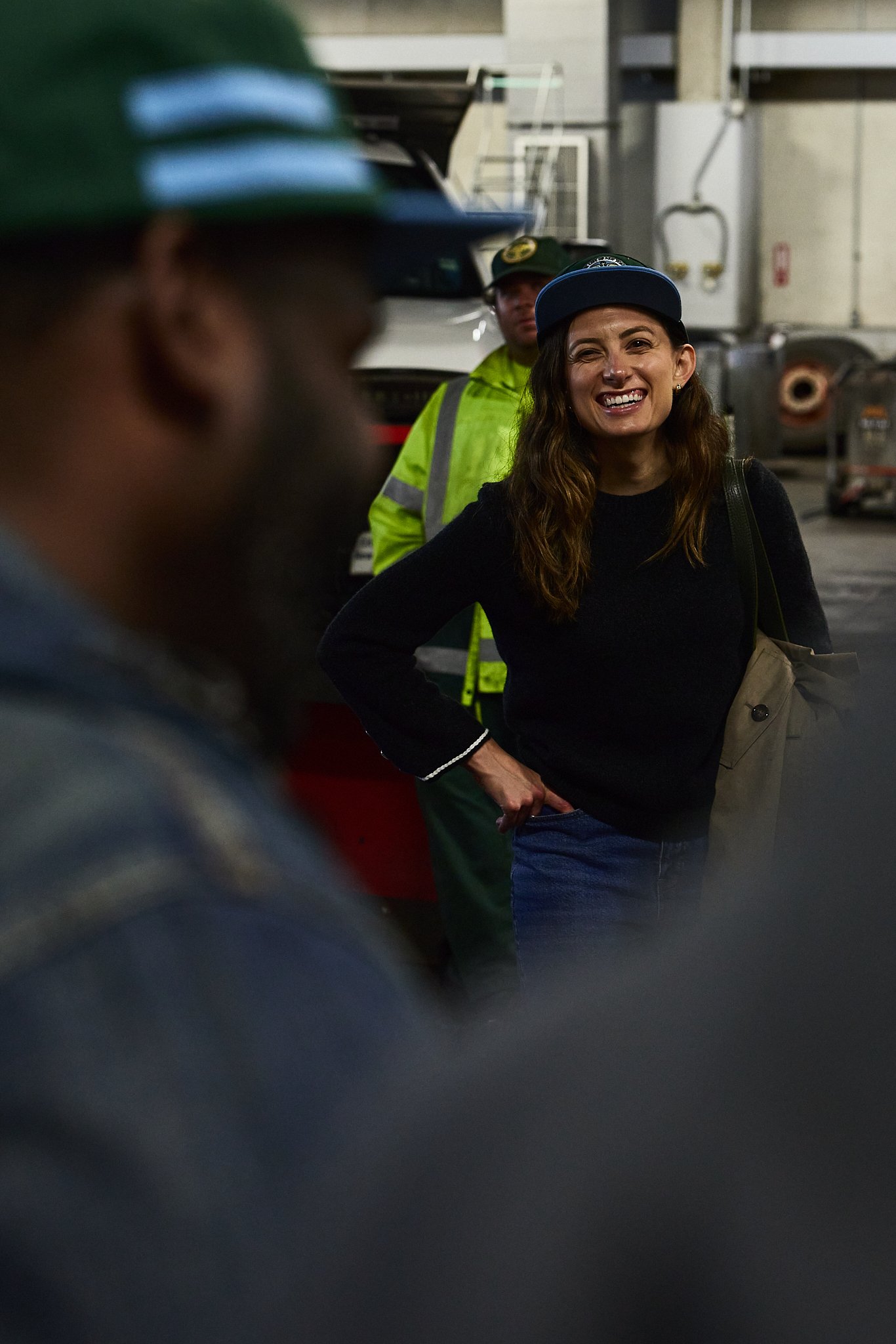 A woman smiling and talking to a person in the foreground inside an industrial or warehouse setting, with a worker in a yellow safety jacket and another worker in a green hat visible in the background.
