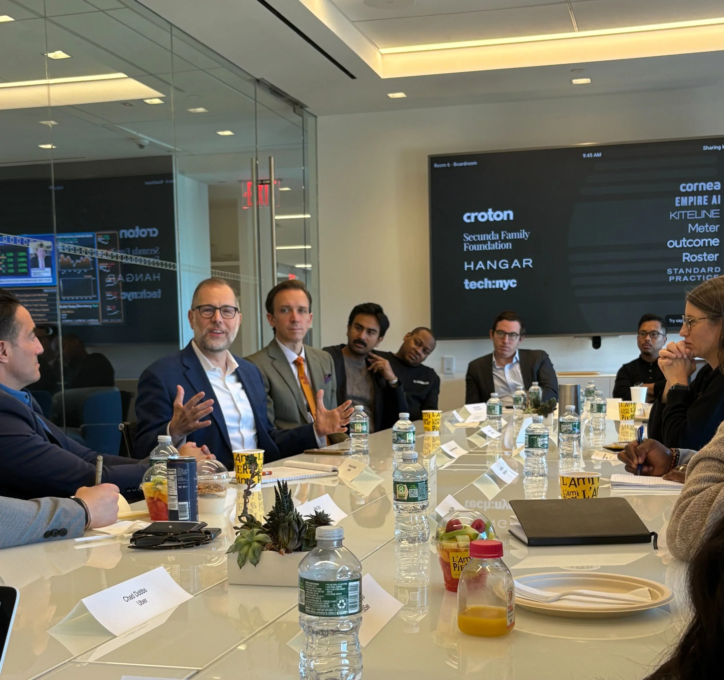 A group of people in business attire sitting around a conference table, engaged in discussion, with water bottles and snacks on the table, and a large screen displaying company names and logos in the background.