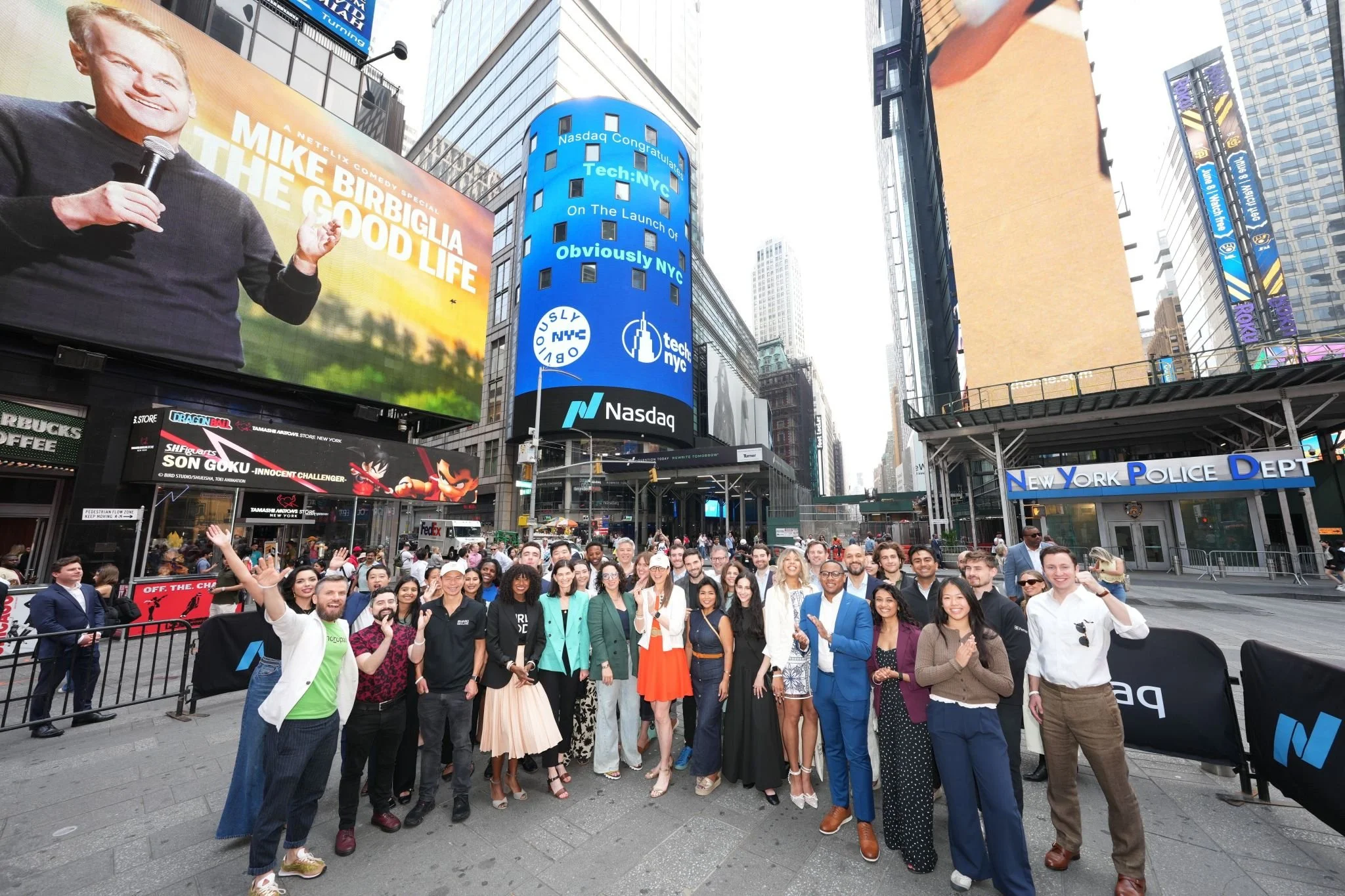 A large group of diverse people posing together outdoors in Times Square, New York City, with digital billboards and buildings in the background.