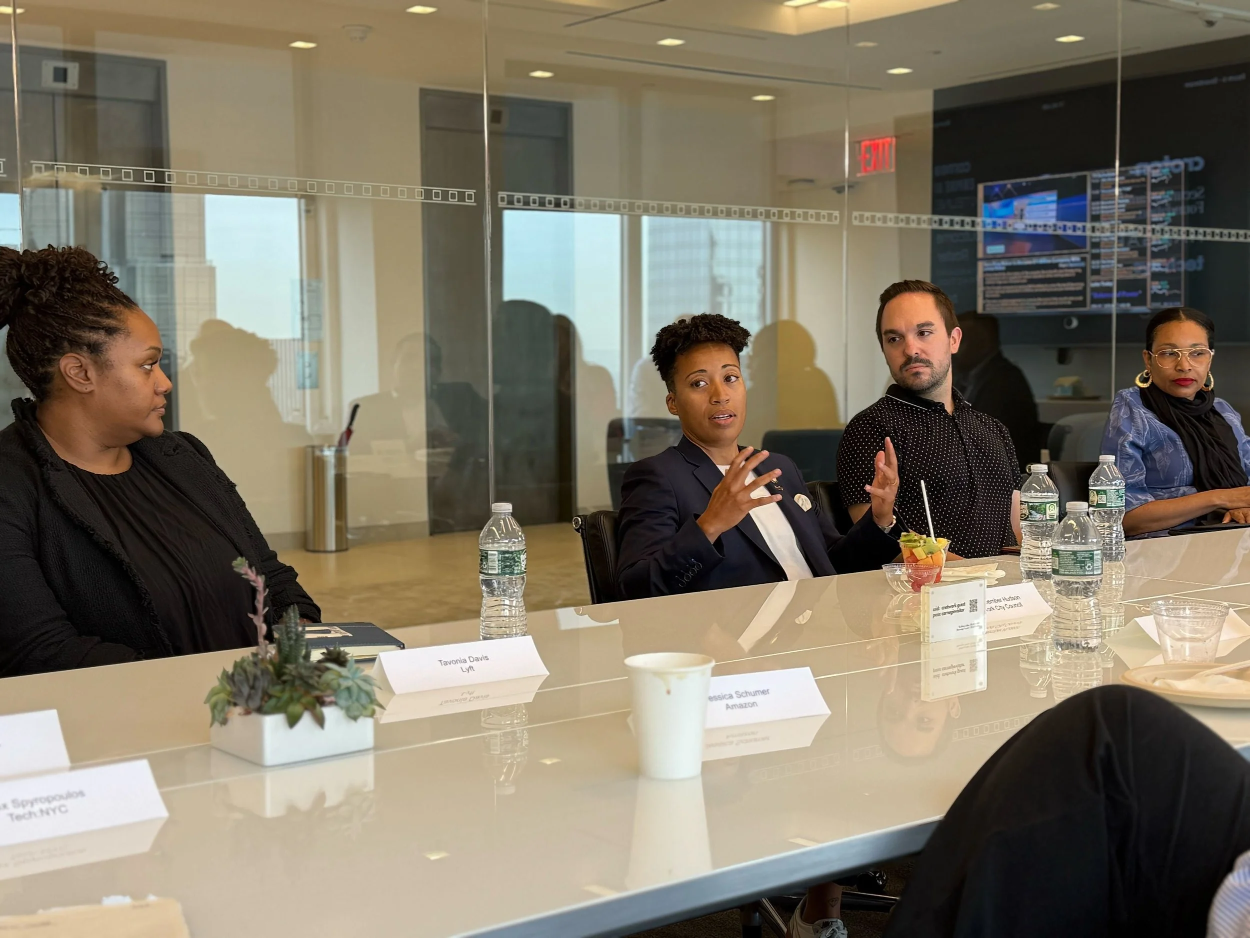 A diverse group of people sitting at a conference table in a modern office, engaged in a discussion. There are water bottles, a small potted plant, and name cards on the table. Four individuals are visible, with one woman speaking, gesturing with her