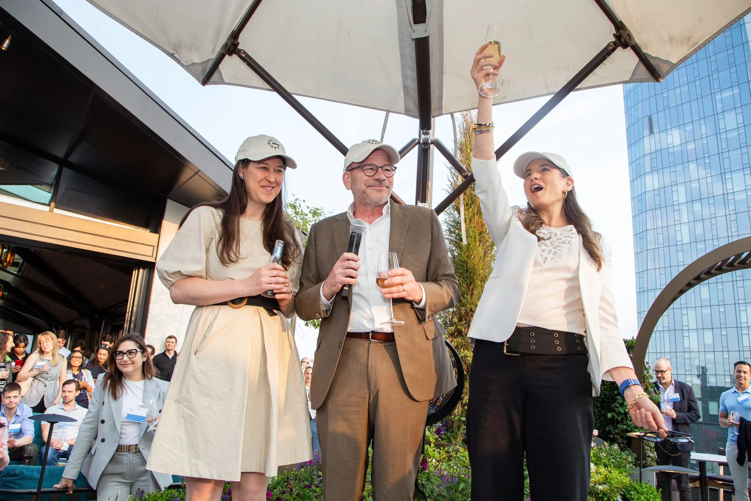 People celebrating outdoors, with three individuals standing under a large umbrella, holding drinks, surrounded by a crowd in a city setting with tall buildings.