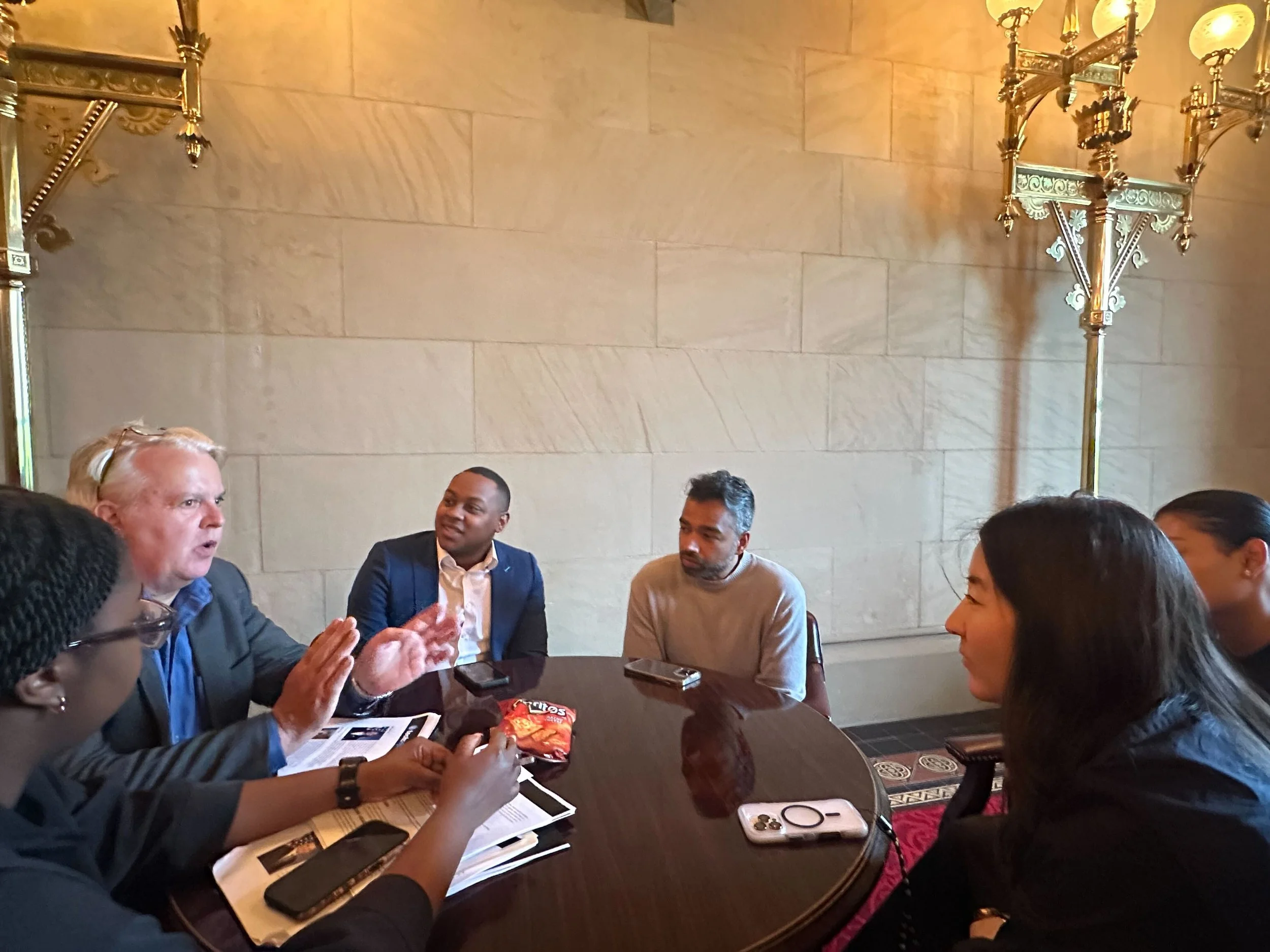 Group of six people sitting around a table in a formal room, engaged in conversation. There are papers, smartphones, and a bag of snacks on the table, and ornate gold light fixtures on the wall behind them.