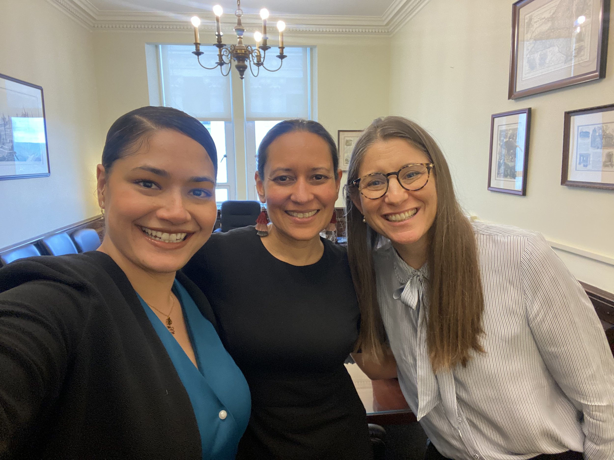 Three women smiling and taking a selfie in a room with framed pictures on the wall.