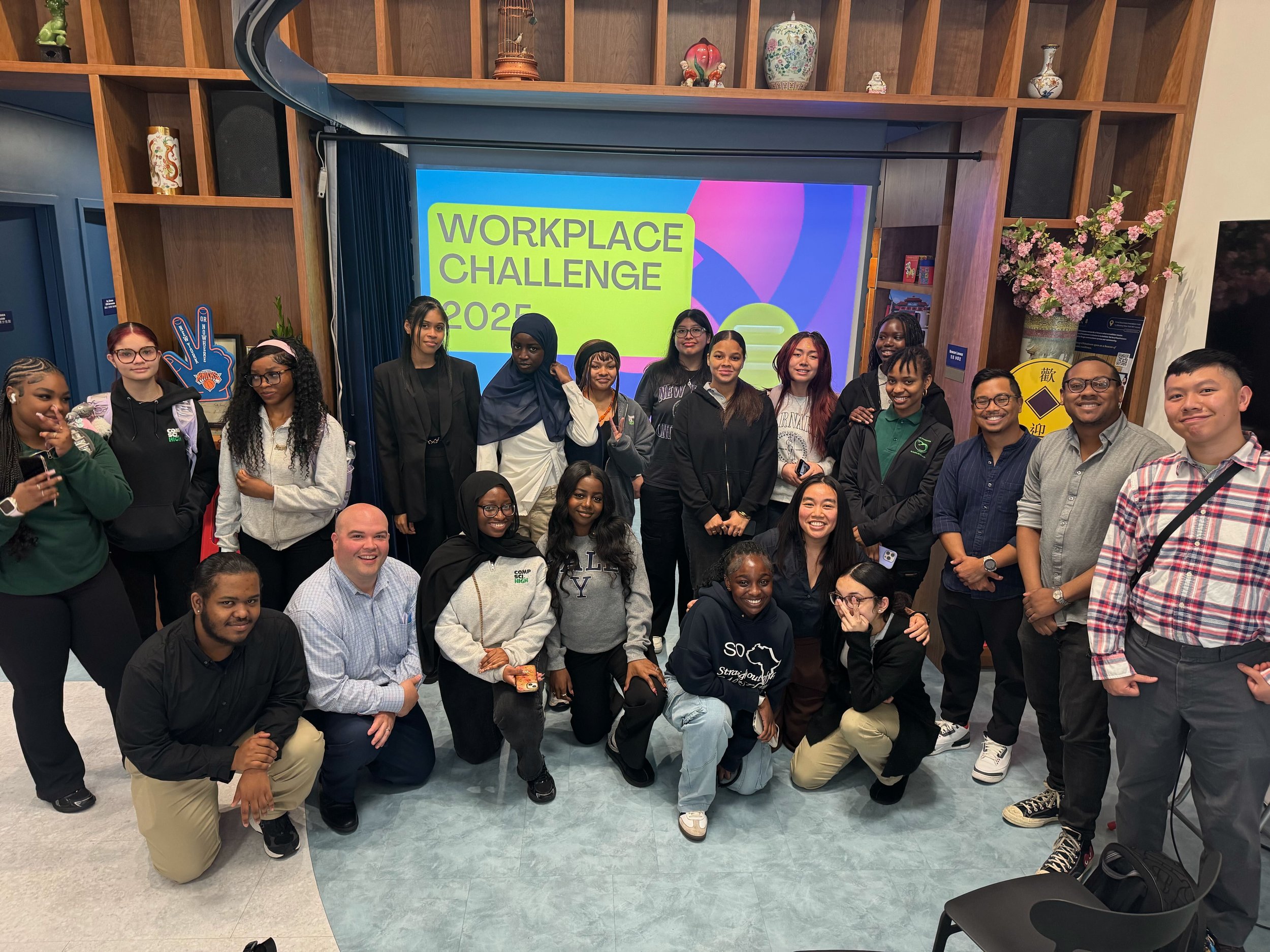 Group of diverse young adults and a few older adults gathered together in a room, smiling for a photo in front of a Signboard that reads "Workplace Challenge 2025." The room is decorated with wooden shelves, vases, and colorful flowers.