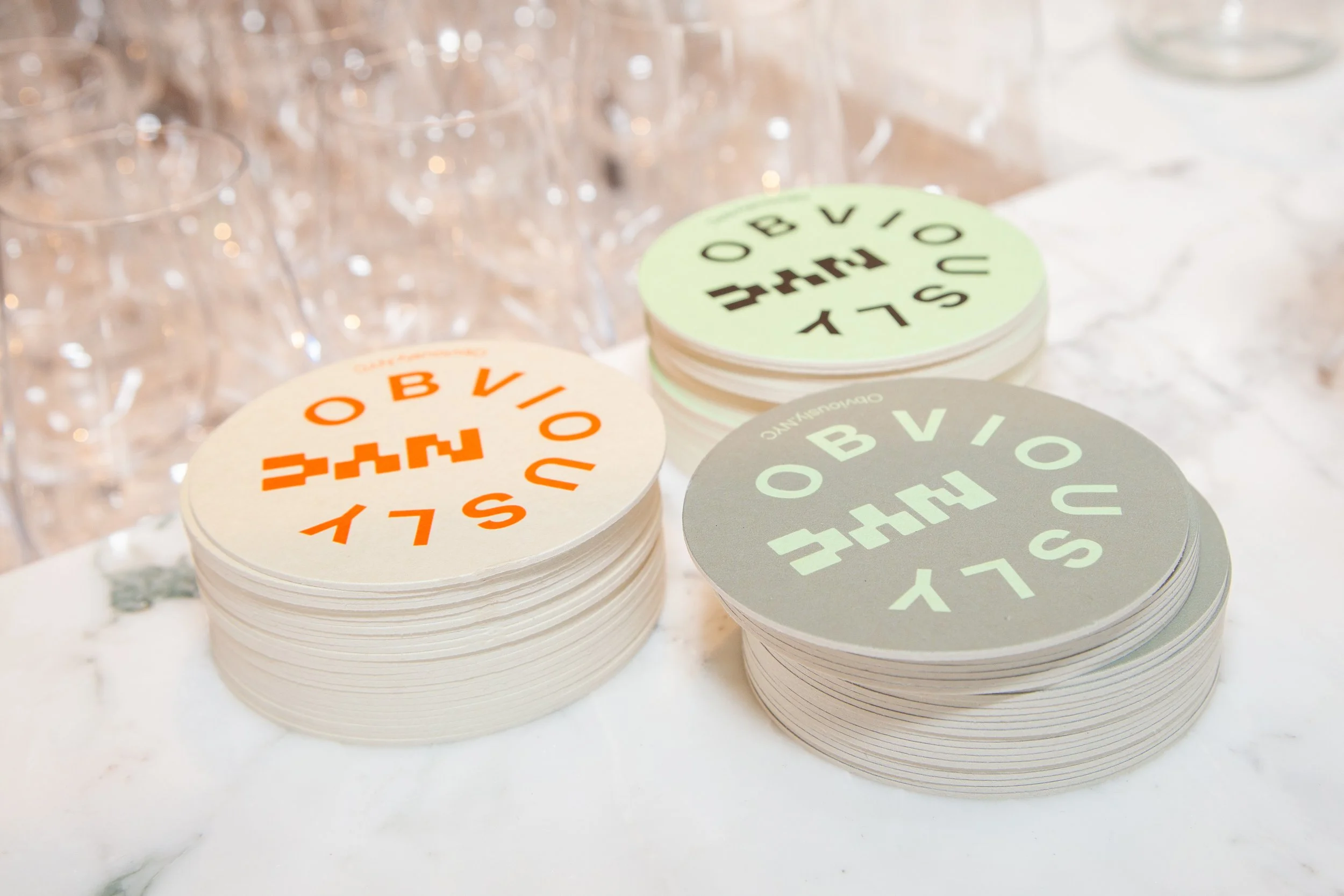 Stacks of coasters with the logo and text 'SUBLIMINAL OBVIOUSE' in different colors on a marble surface, with empty glasses in the background.