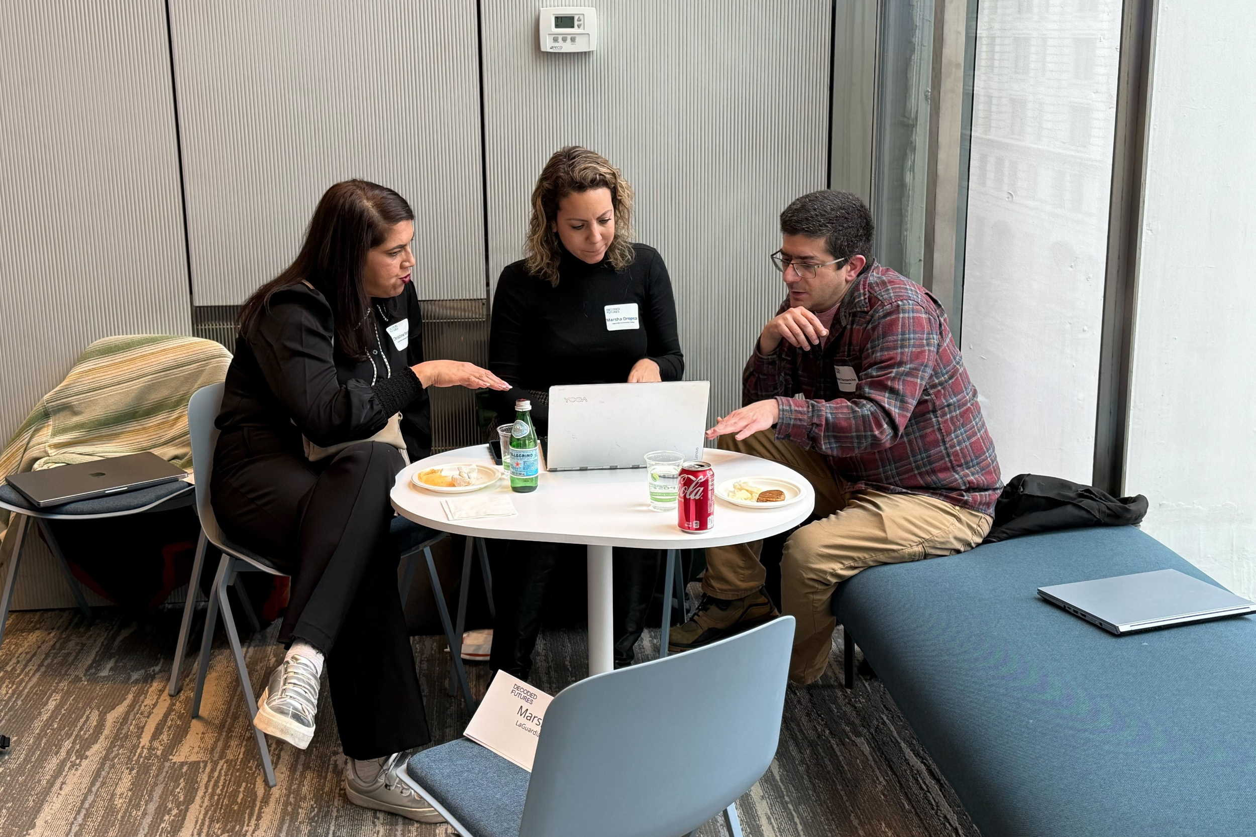 Three people sitting around a small round table, engaged in a discussion, with laptops and drinks in front of them, in a modern office space.