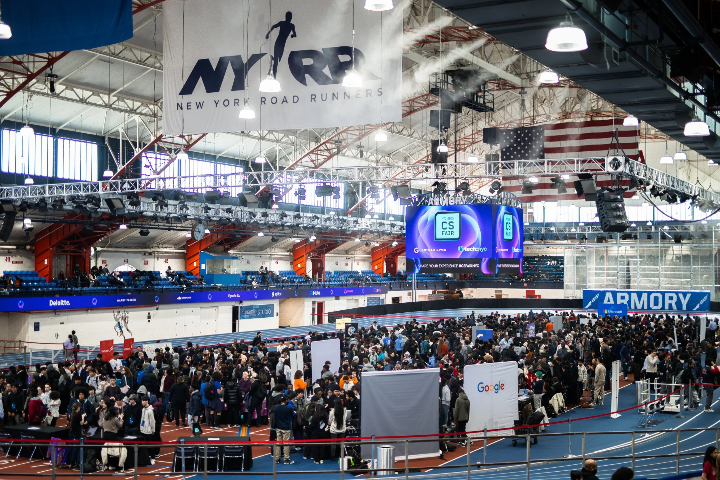 Crowd of people at a tech conference in an indoor stadium with banners reading "New York Road Runners" and "Armory." Large digital screens display event information, and attendees are gathered around booths, with some walking along the tracks.