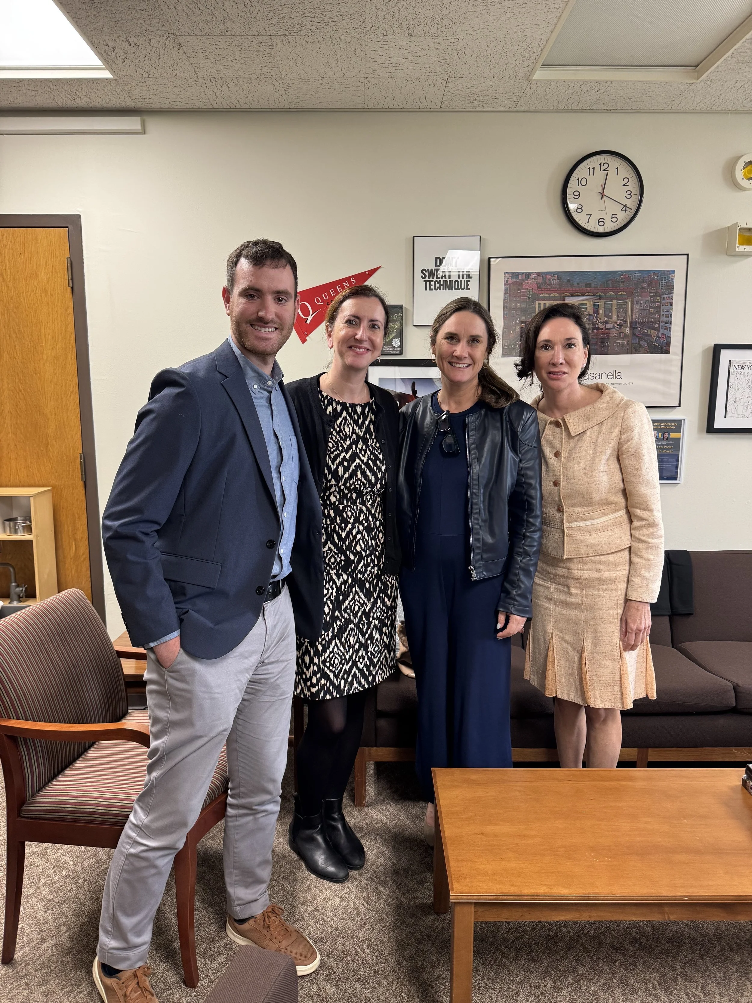Four people standing together in an office, smiling at the camera. The background shows framed pictures, a wall clock, and a desk.