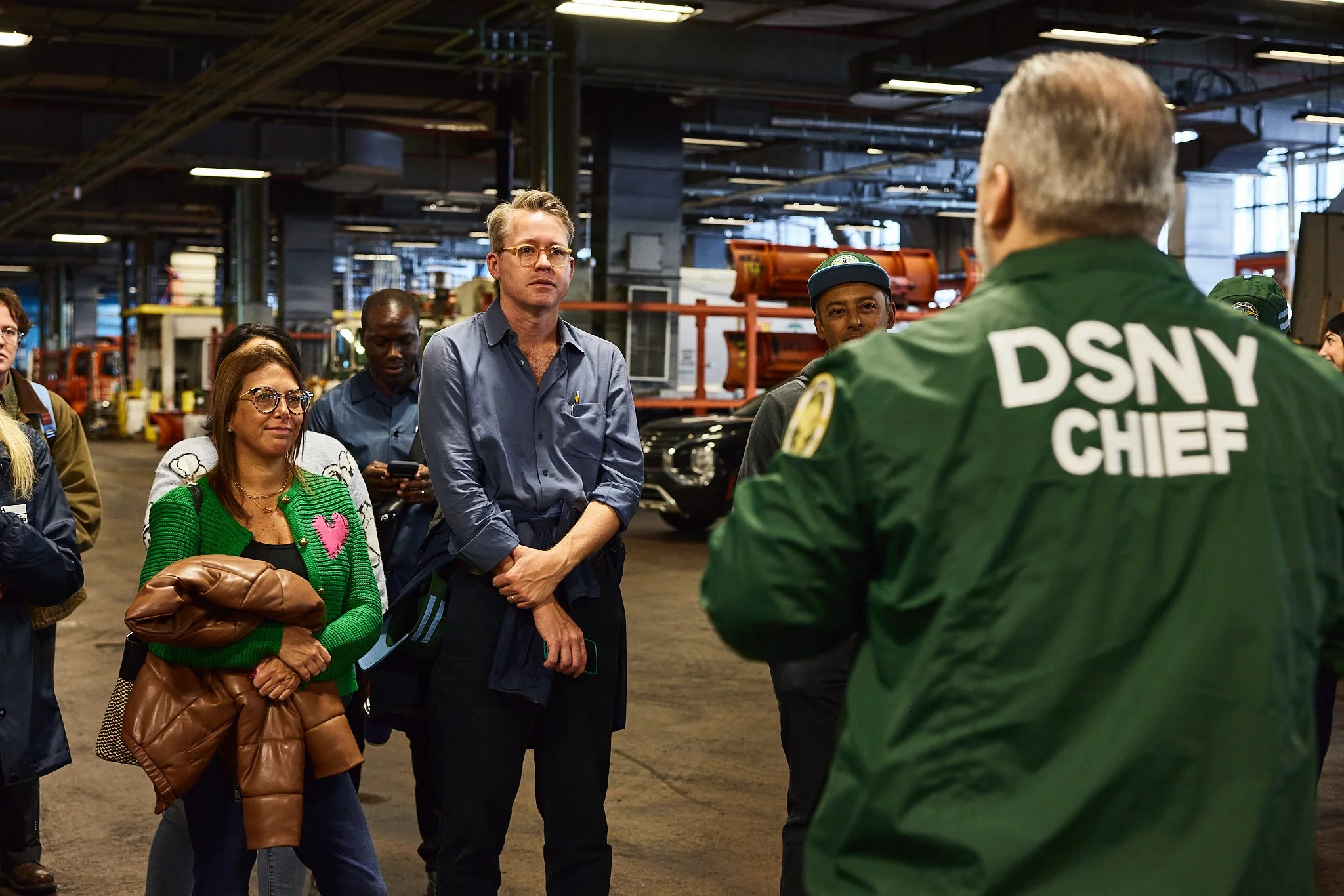 A group of people listening to a person with a DSNY CHIEF jacket, inside a warehouse with equipment and cars.