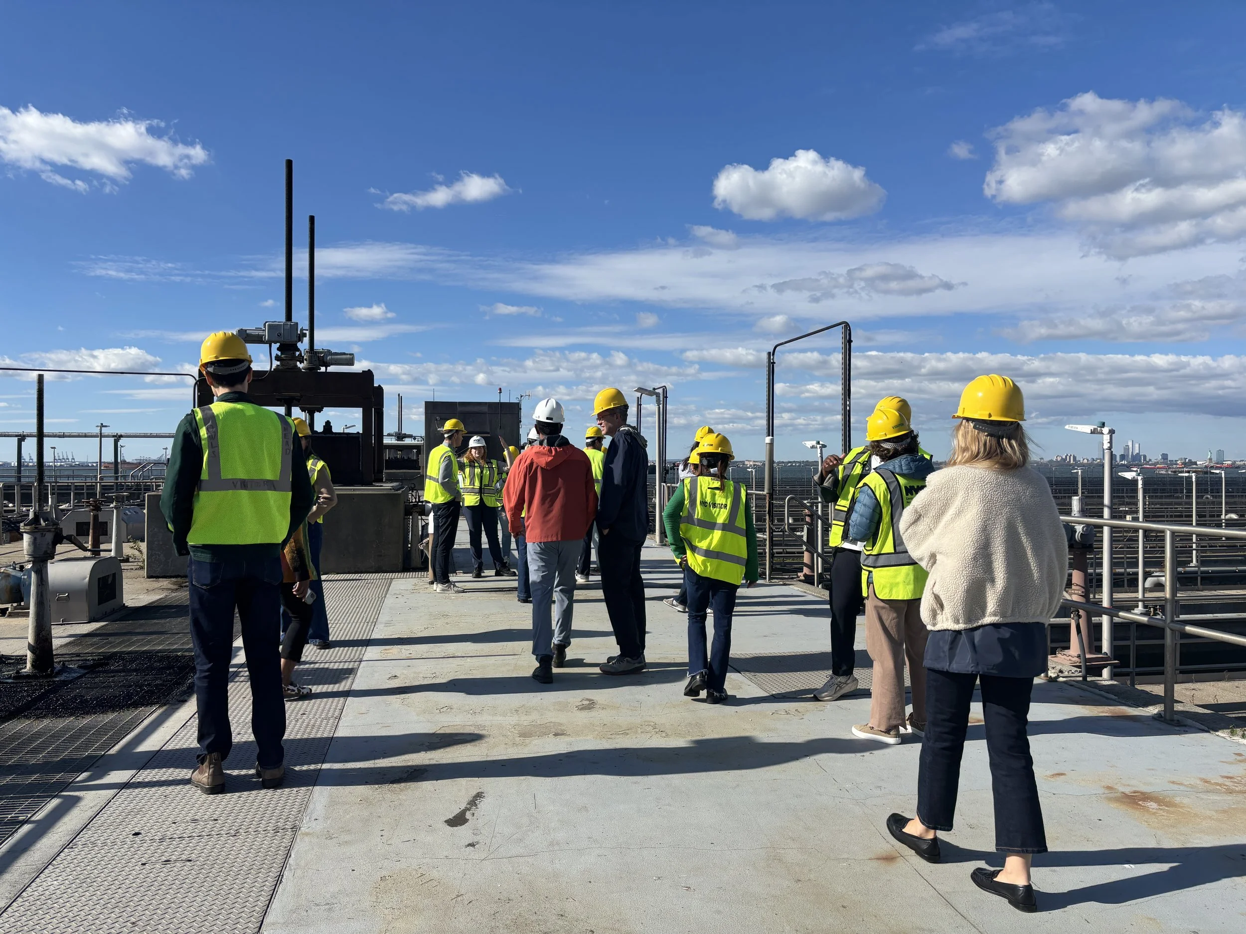 Group of people wearing yellow and white safety helmets and reflective vests on a rooftop industrial site with metal railings, pipes, and a city skyline in the distance under a partly cloudy sky.