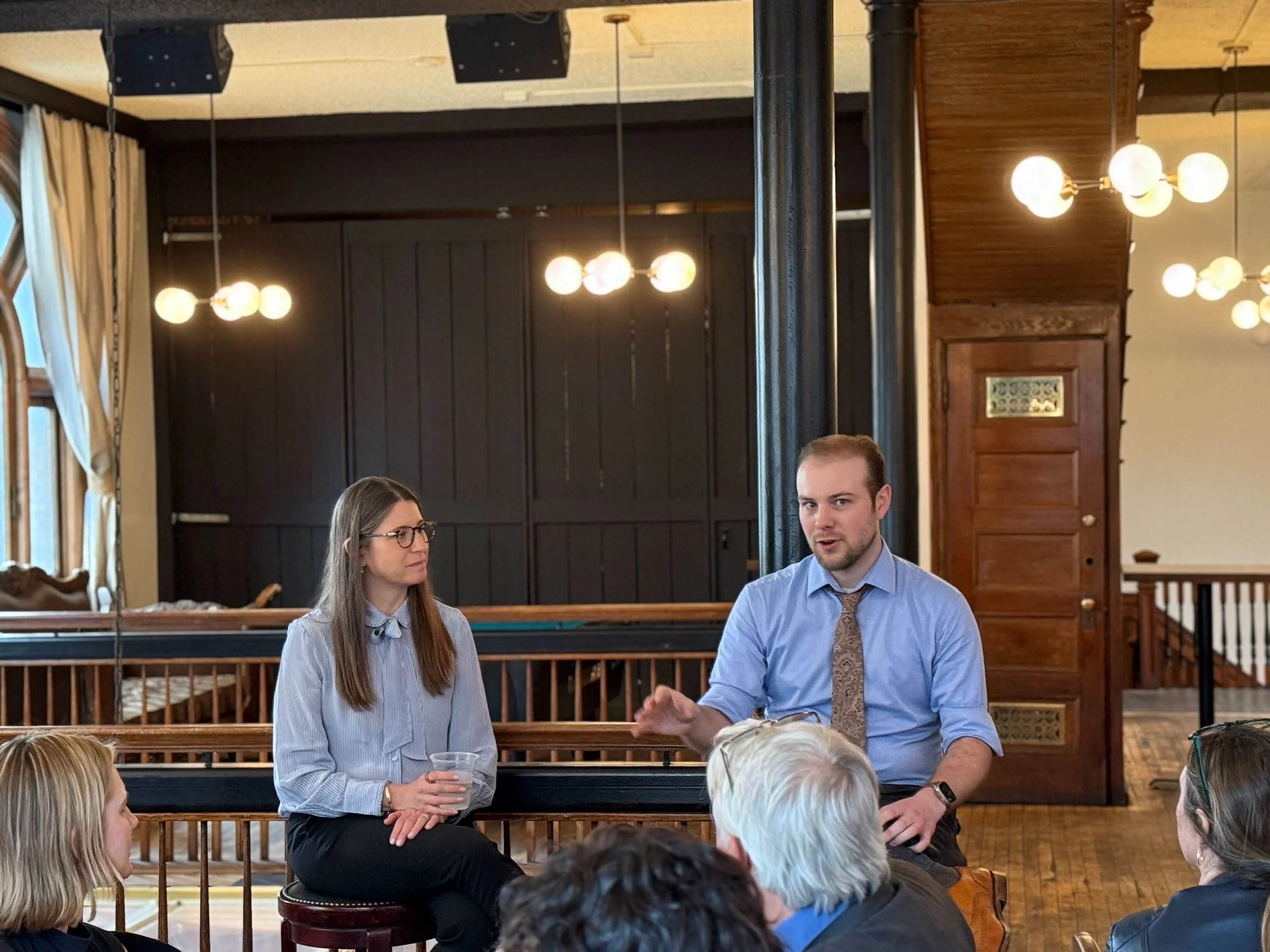A man and a woman are seated and speaking to a group of people in a room with wooden paneling, chandeliers, and large windows with curtains.