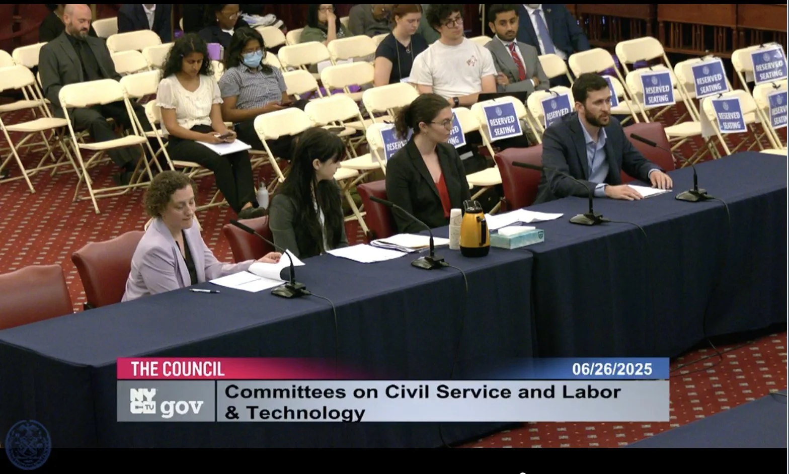 People attending a formal government committee hearing with reserved seating signs, sitting at a long conference table with microphones, documents, and a yellow thermos, with some attendees using phones and others taking notes, in a wood-paneled room