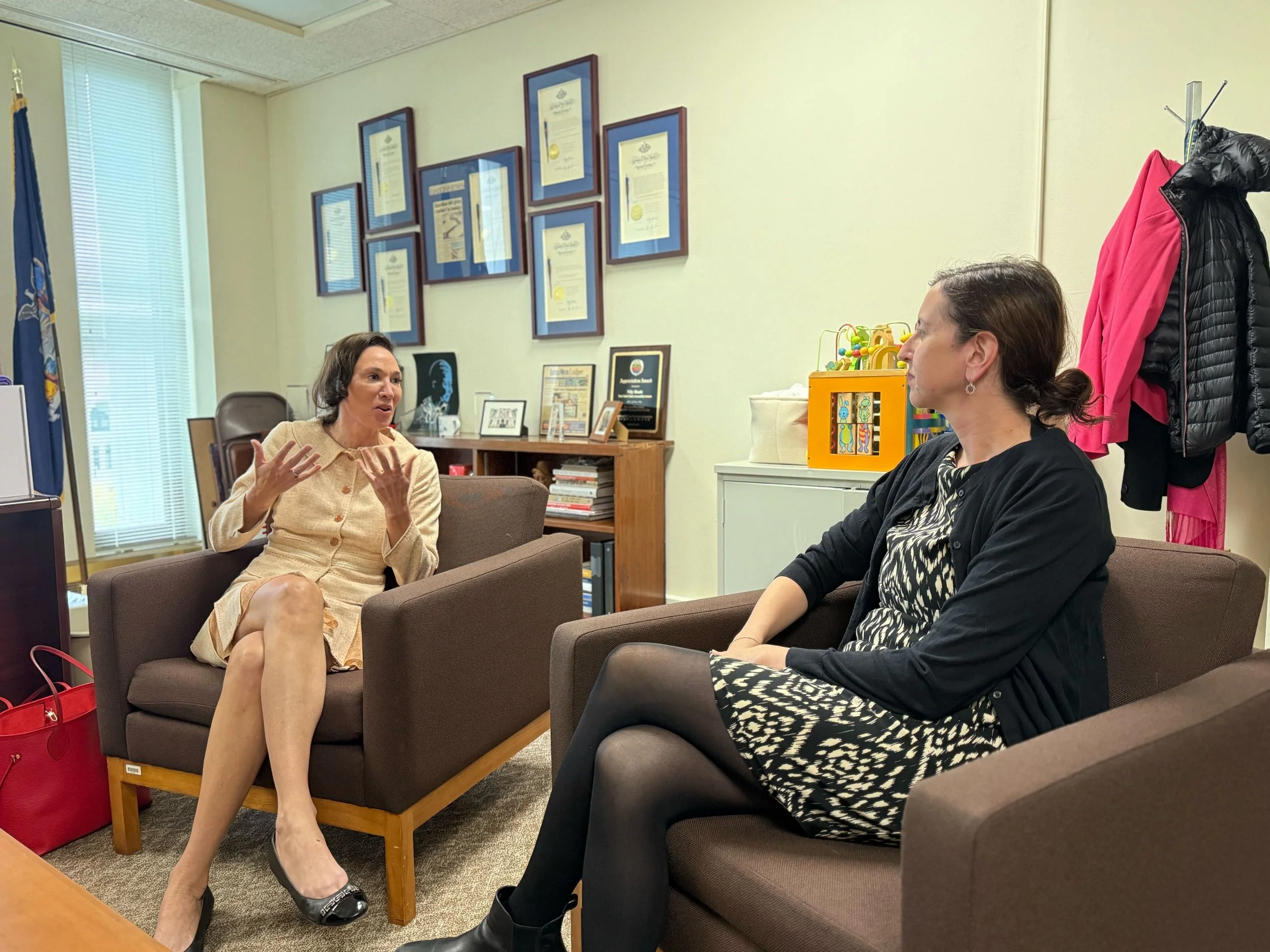 Two women sitting on brown armchairs in an office, engaged in conversation. One woman is gesturing with her hands, wearing a beige dress, while the other is listening, wearing a black and white dress with a black cardigan. There are framed certificat