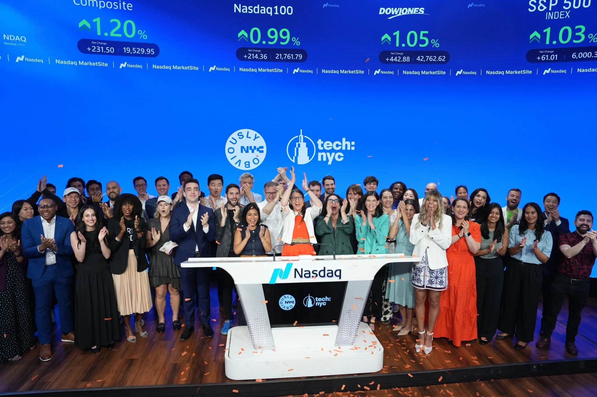 A large group of people gathered on stage at a Nasdaq event, celebrating with smiles, claps, and confetti. The backdrop displays stock indices and logos for Nasdaq and Tech: NYC.