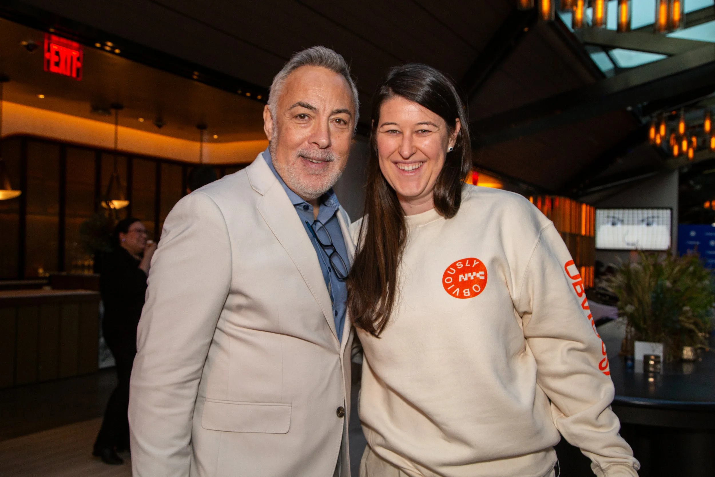A man and a woman smiling and posing together indoors; the woman is wearing a beige sweatshirt with orange text and graphics, the man is dressed in a light-colored blazer and blue shirt.