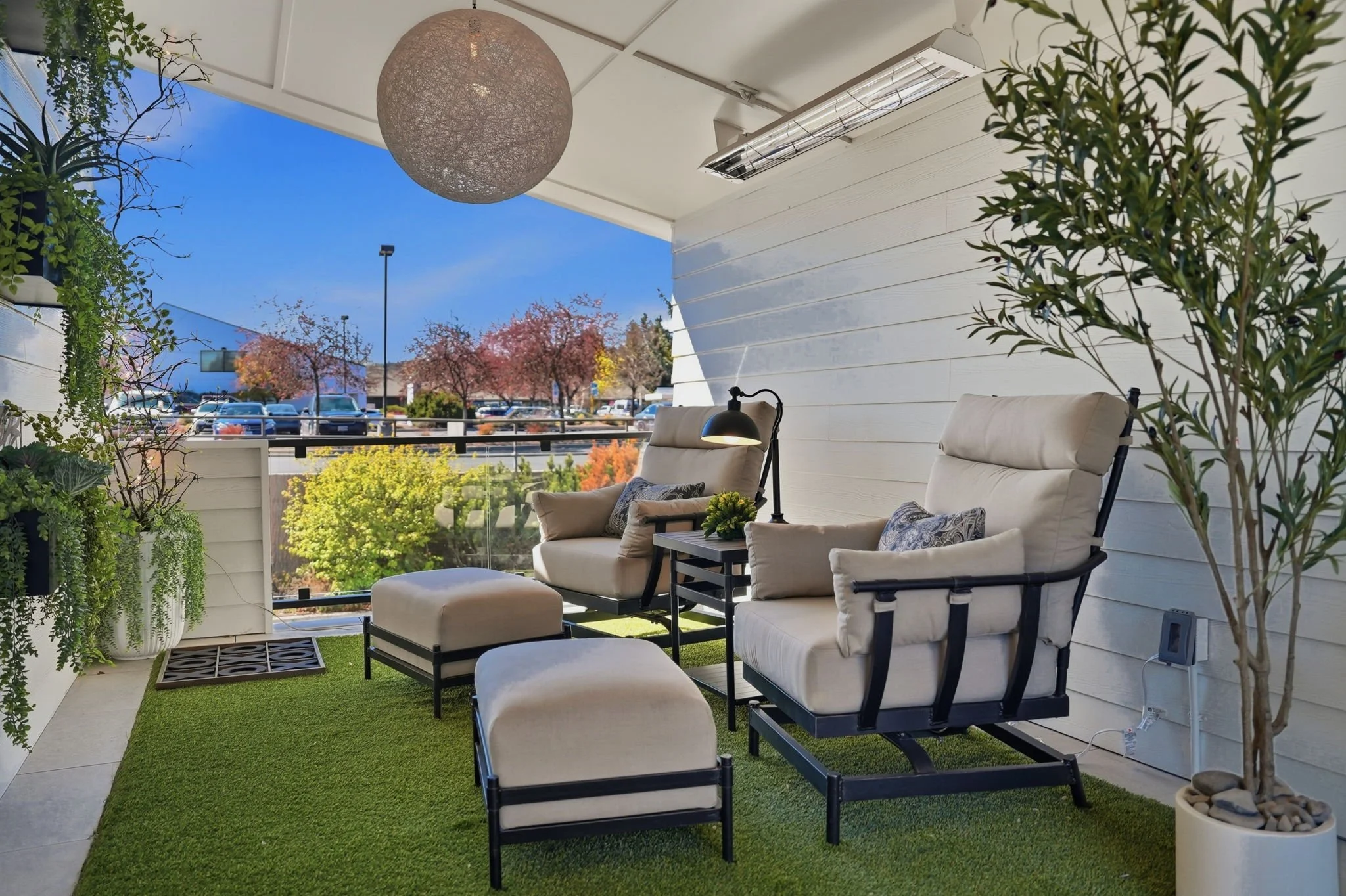 A cozy outdoor balcony with white furniture, including armchairs with cushions, a small table with a potted plant, and decorative pillows. There are lush green plants in white pots along the edges, a green artificial grass rug, and a view of a parking lot with trees and colorful foliage outside. The ceiling has a spherical hanging light and an air conditioning unit.