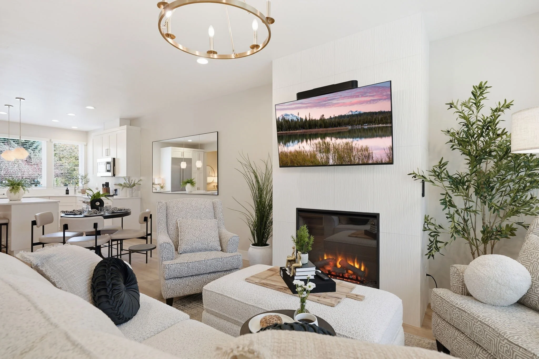 A modern living room with white walls, a flat-screen TV above a fireplace, and various patterned chairs and sofas. There are plants, a floor lamp, and a wooden coffee table with decorative items. The room is well-lit with natural and artificial light, and visible kitchen and dining area in the background.