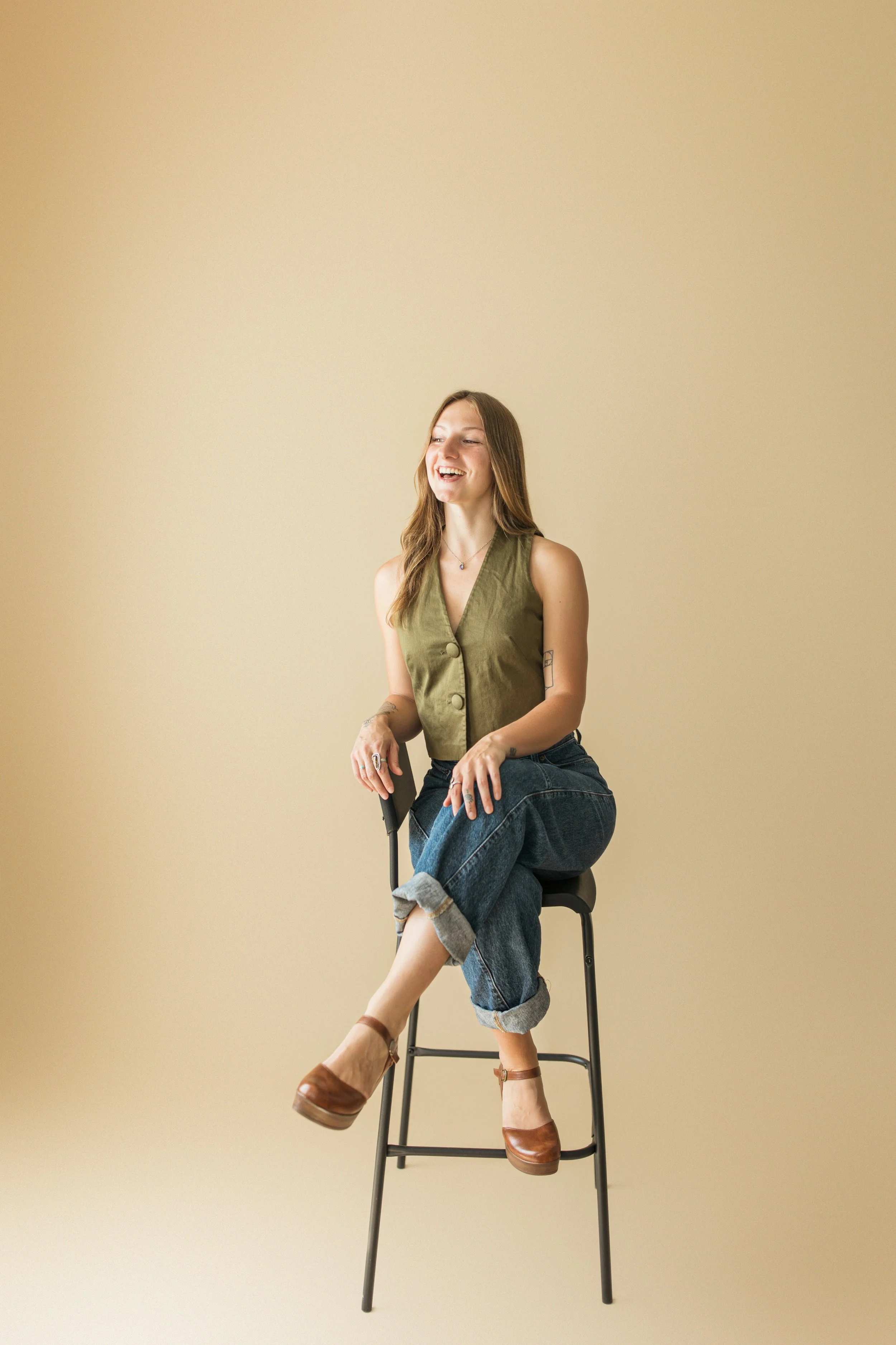 A young woman with long hair smiling while sitting on a high stool against a plain beige background. She's wearing a sleeveless green vest, dark cuffed jeans, and brown platform shoes, with visible tattoos and rings on her fingers.