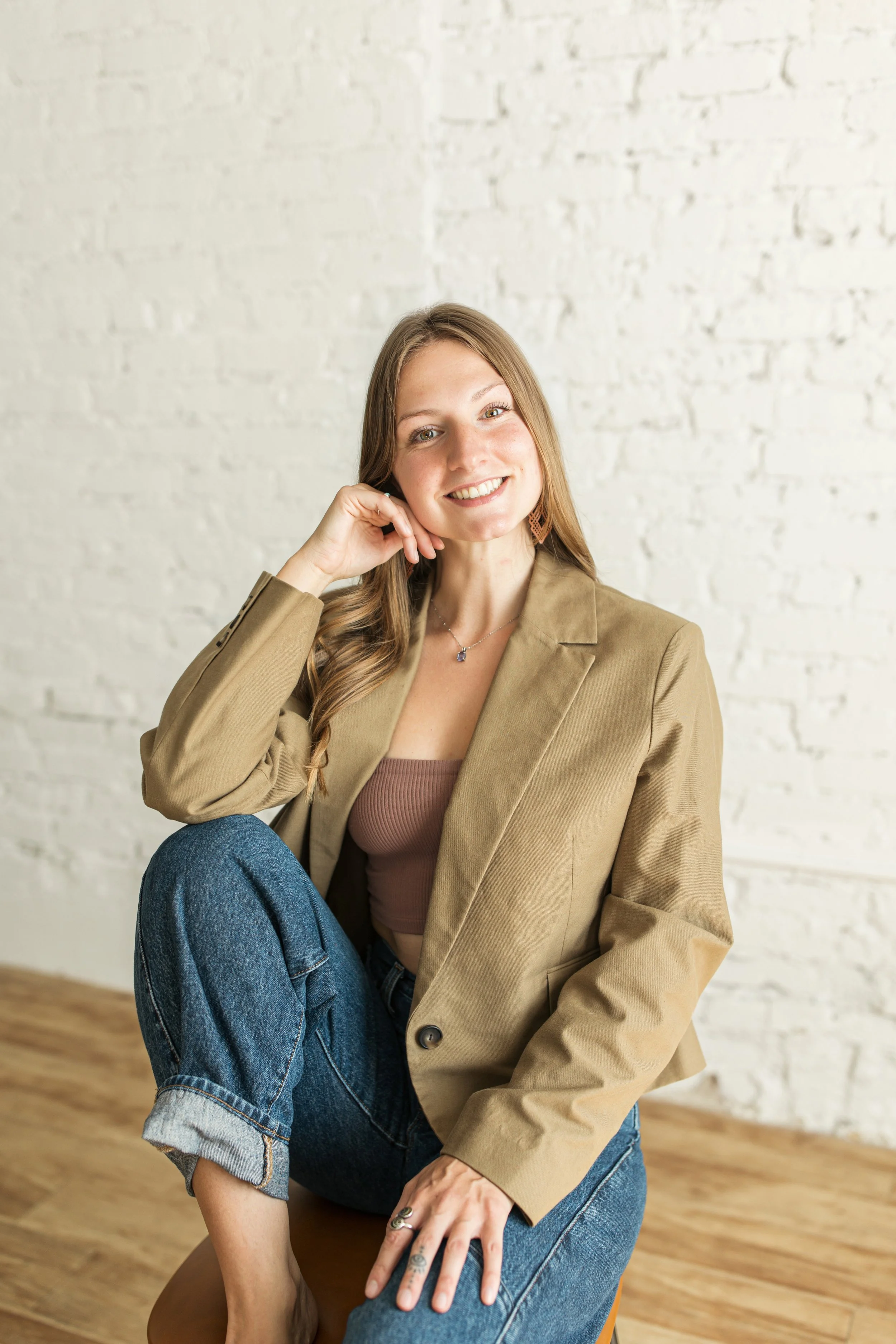 A young woman with long, wavy brown hair, wearing a beige blazer over a mauve top and blue jeans, sitting on a wooden chair in front of a white brick wall, smiling at the camera.