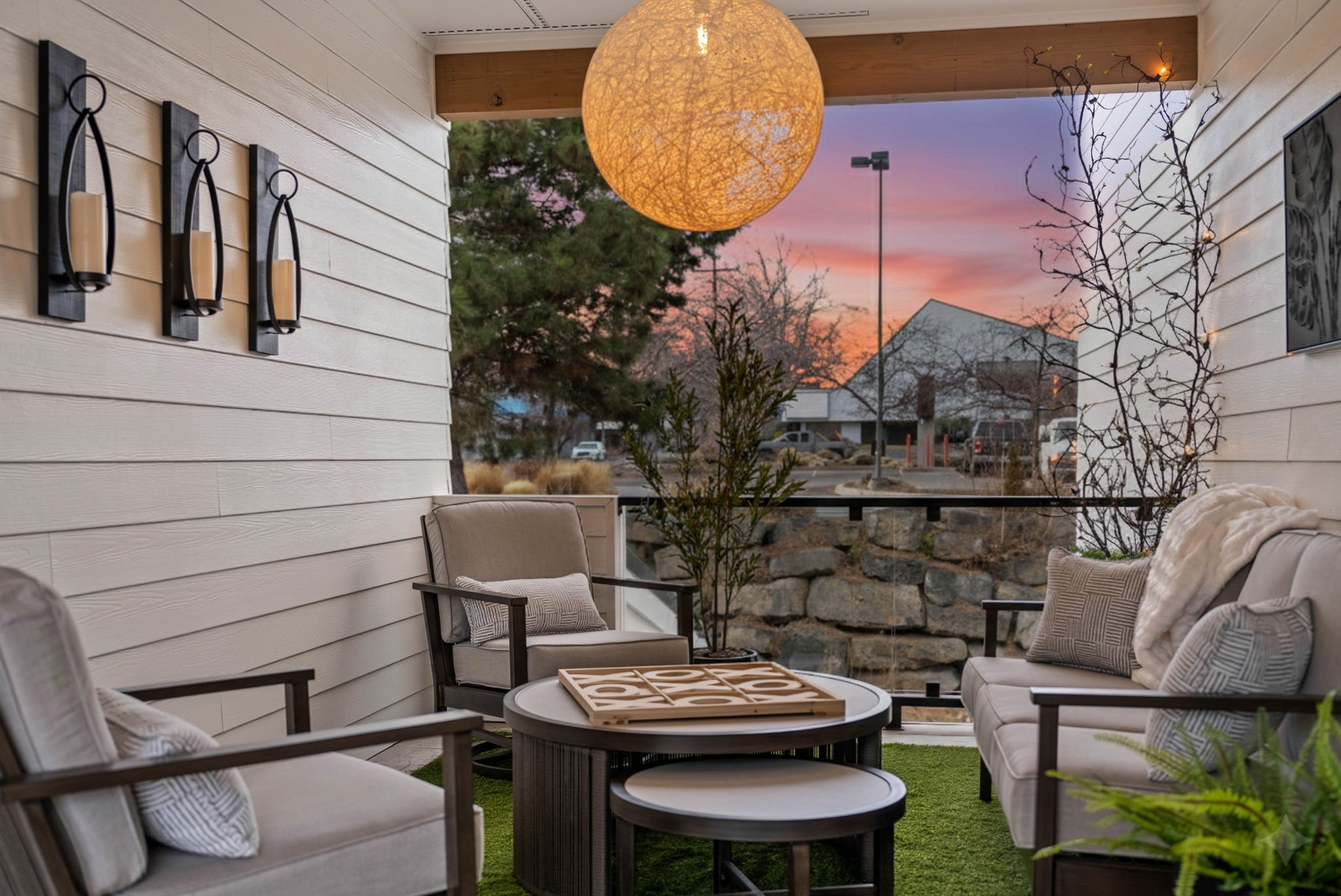 Outdoor porch seating area with beige cushioned chairs, a round table, and decorative pillows. A large spherical hanging light and string lights create a cozy atmosphere, with trees and houses visible outside during sunset.