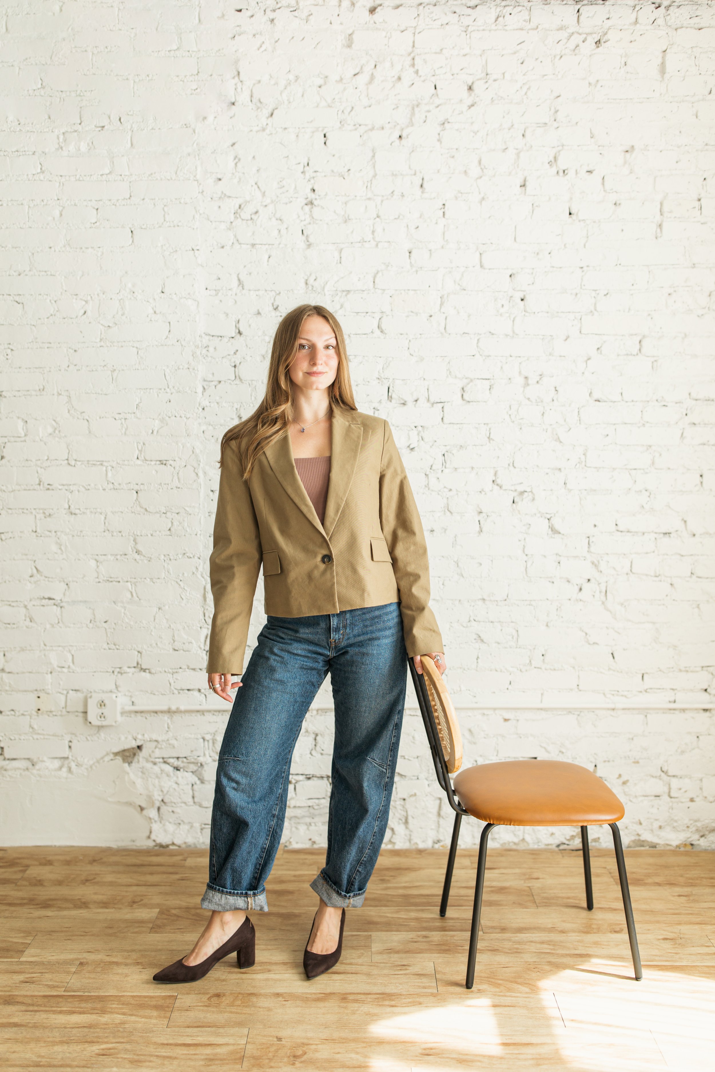 A woman with long hair wearing a beige blazer, a pink top, and rolled-up jeans standing next to a chair in front of a white brick wall.