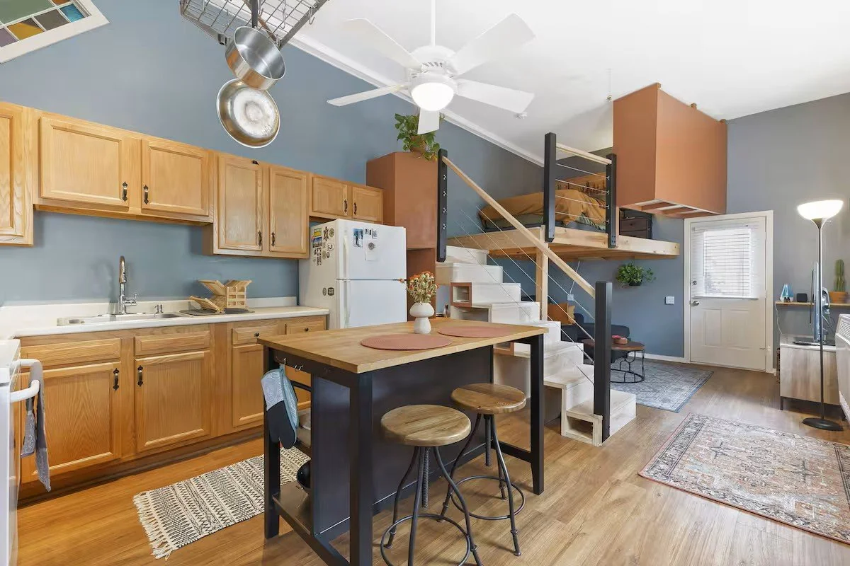 Open-concept kitchen and living area with wooden cabinets, white appliances, a small dining table with two stools, staircase to a lofted bedroom, and various decorative elements.