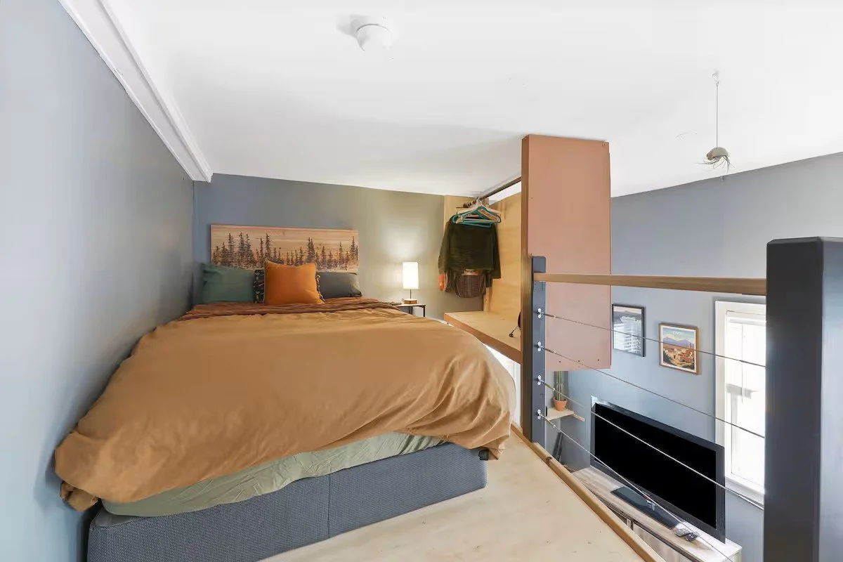 A small bedroom with a bed against a blue wall, a nightstand with a lamp, an open closet with hanging clothes, and a TV on a stand below framed pictures on the wall.