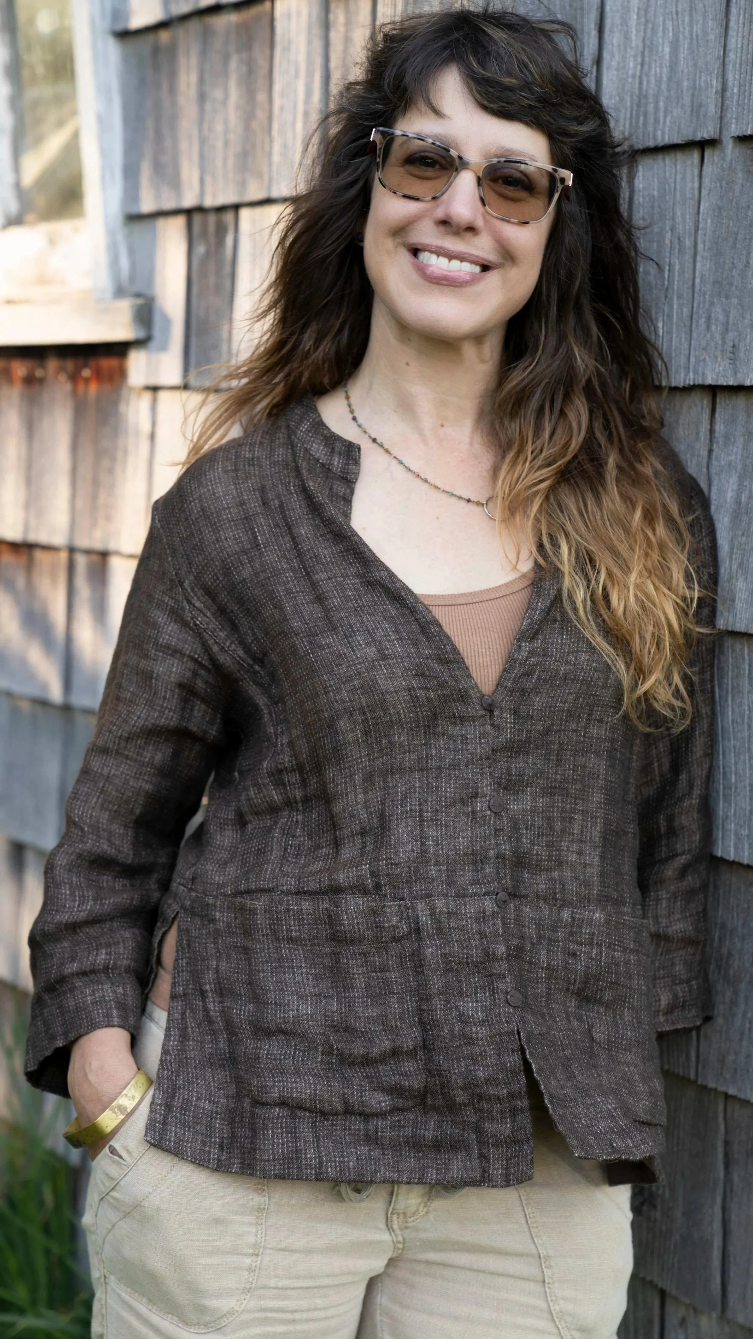 Natalia Farber - lead garden designer for Gardenwise, smiling - standing against a weathered wooden wall, wearing a brown button-up jacket over a tan top, with a beaded necklace, gold bracelet, and beige pants.