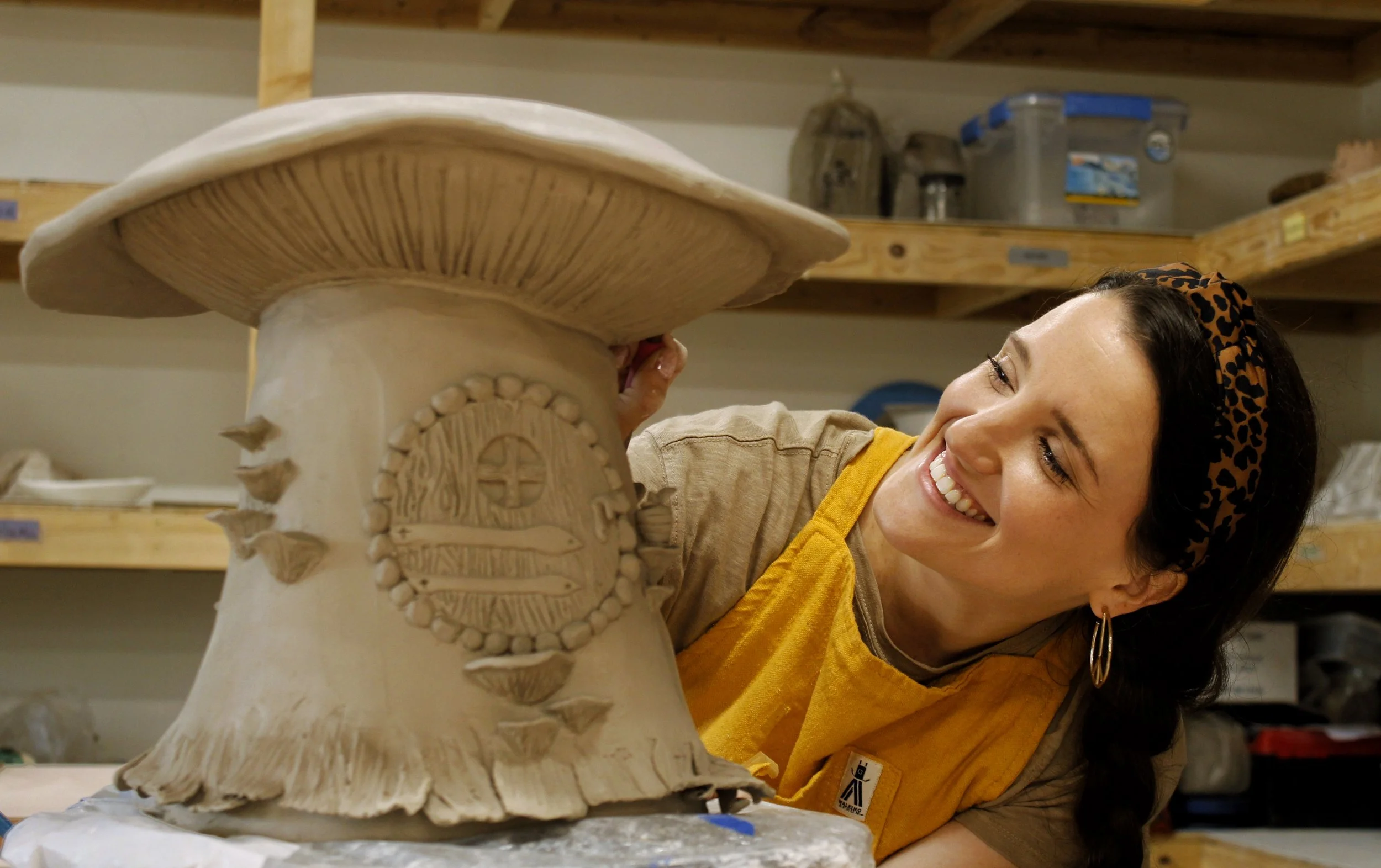 A woman wearing a leopard print headband and a beige and yellow apron smiles while working on a ceramic sculpture in a pottery studio. The sculpture features a large, detailed mushroom with decorative elements on its base.