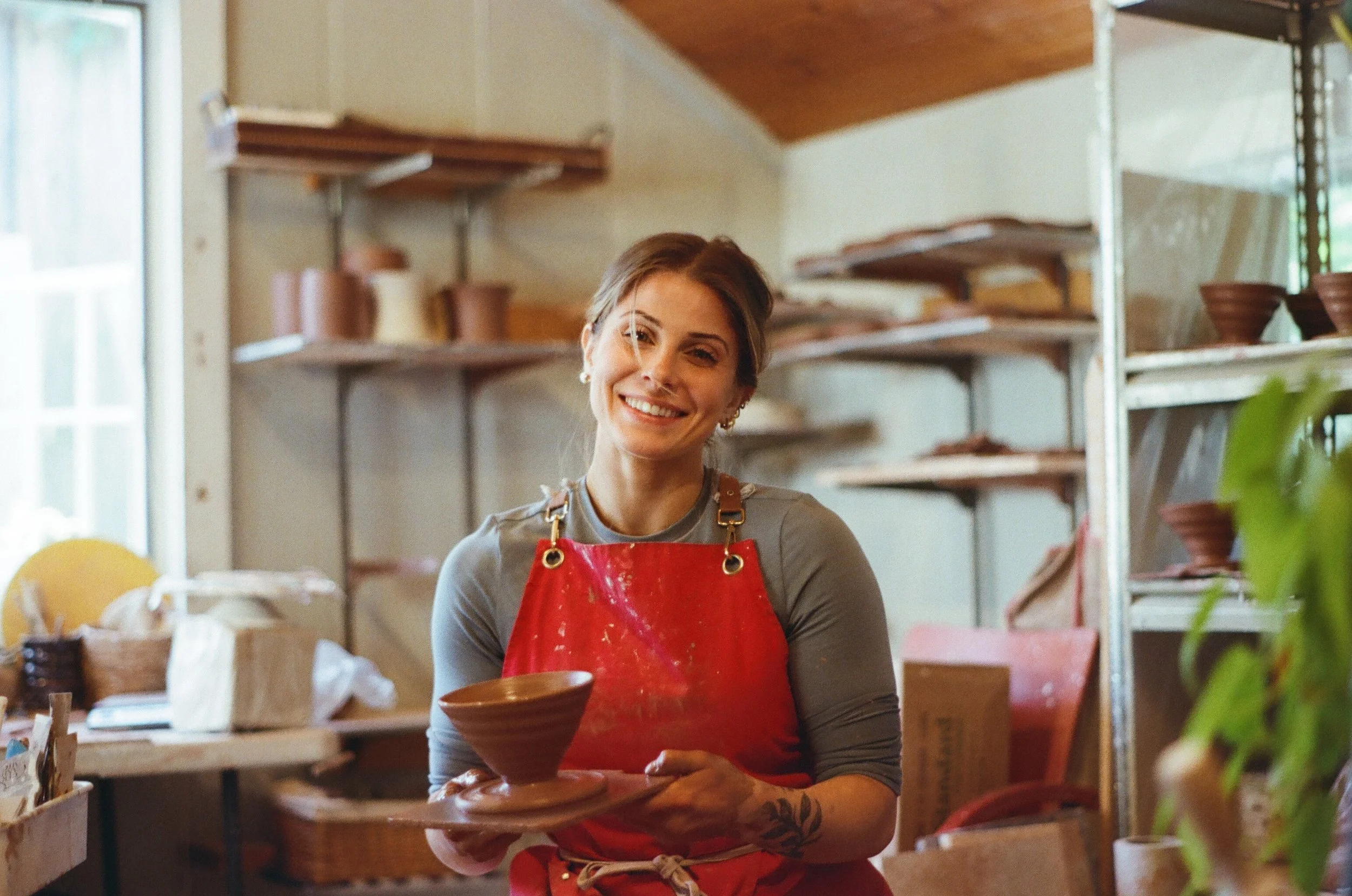 A smiling woman in a pottery studio wearing a red apron holding a brown ceramic bowl, with shelves full of pottery items in the background.