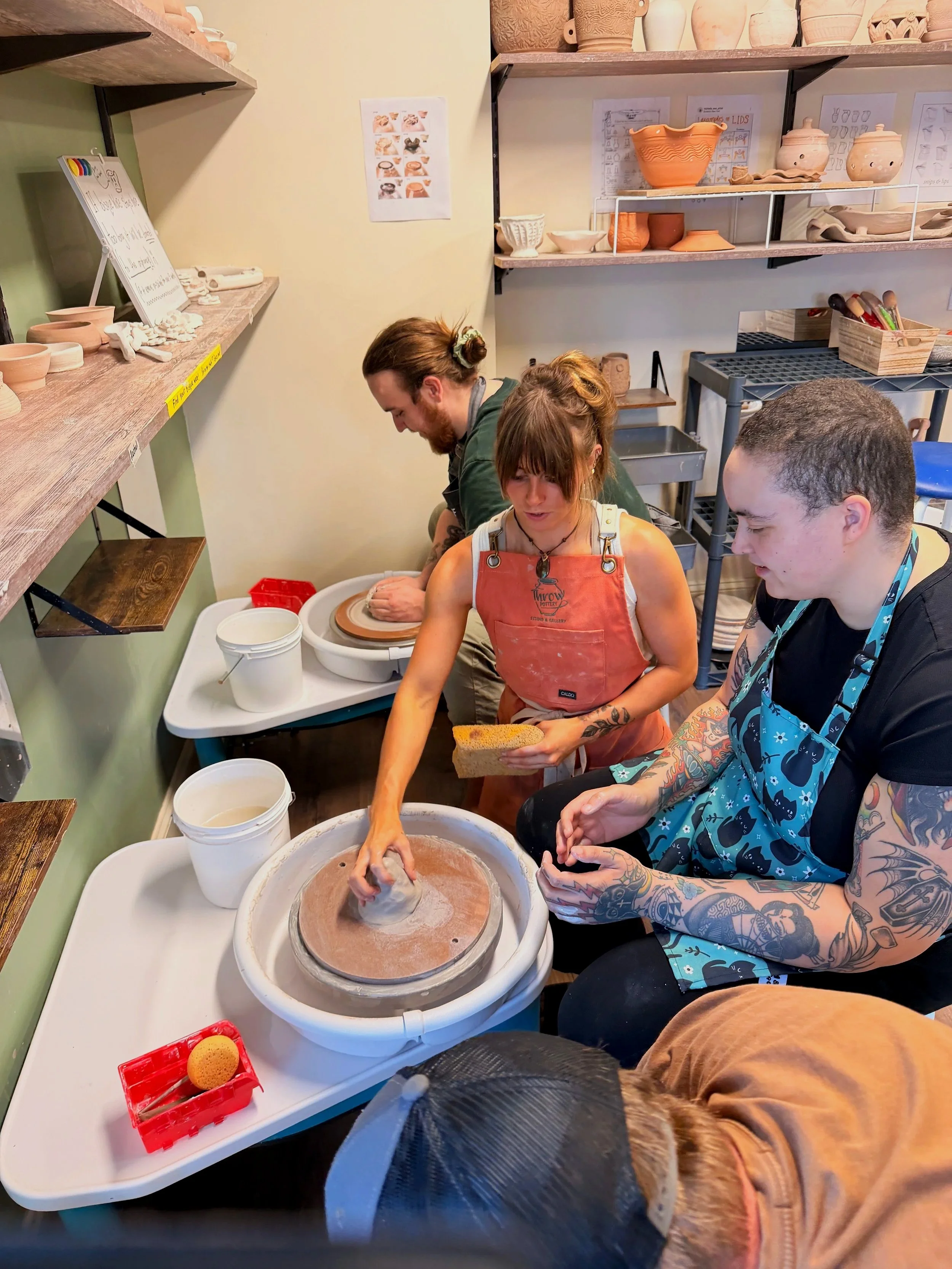 Three people, including a woman with tattoos, are working on pottery-making at a ceramics studio. The woman is shaping a clay piece on a pottery wheel, with two men assisting or observing. The workspace has shelves with finished and unfinished pottery, containers of water and clay, and pottery tools.