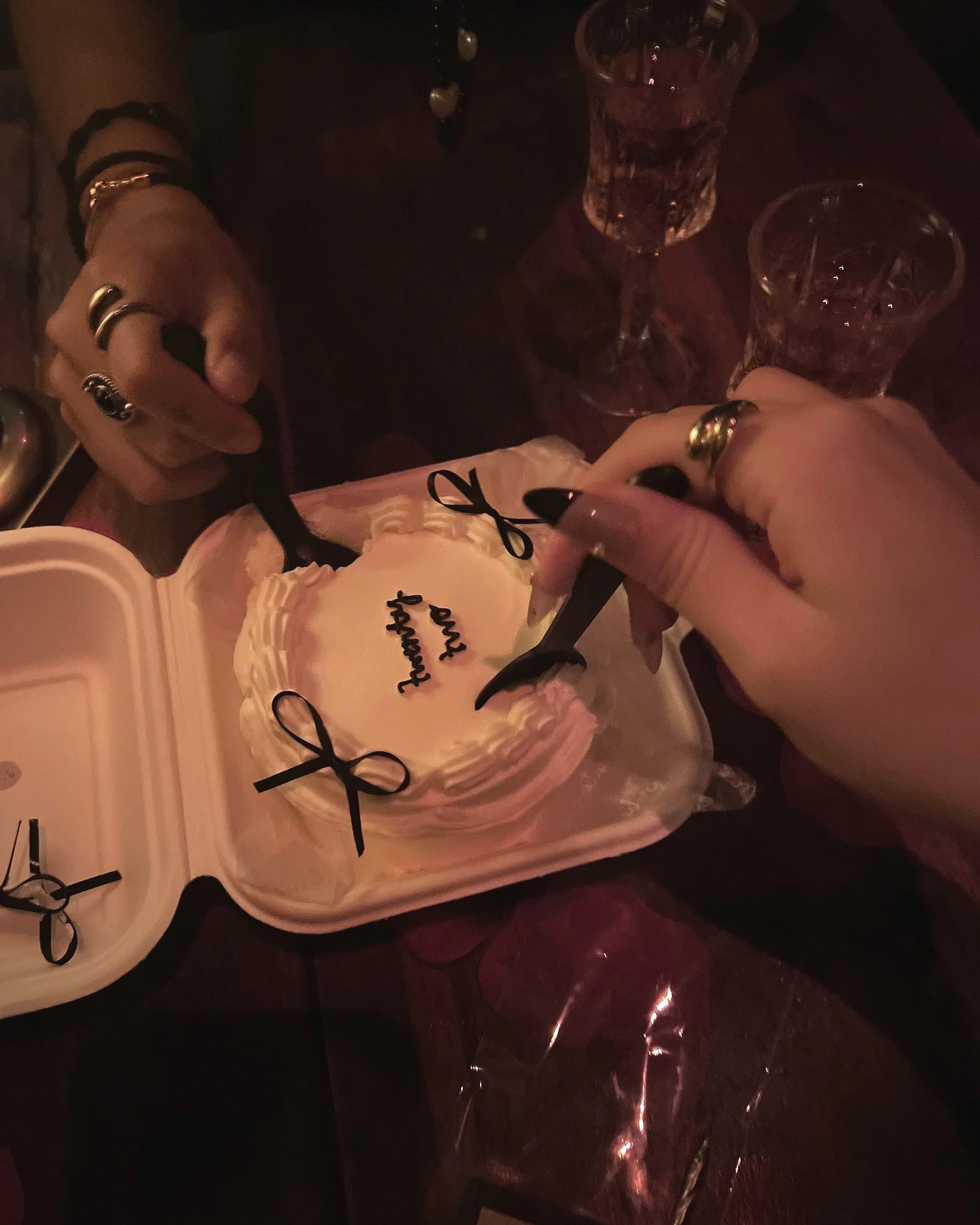 Two people are cutting a heart-shaped birthday cake with 'Happy Birthday' written on it in black icing. There are two glasses of what appears to be pink drinks on the table, and both individuals are wearing rings.