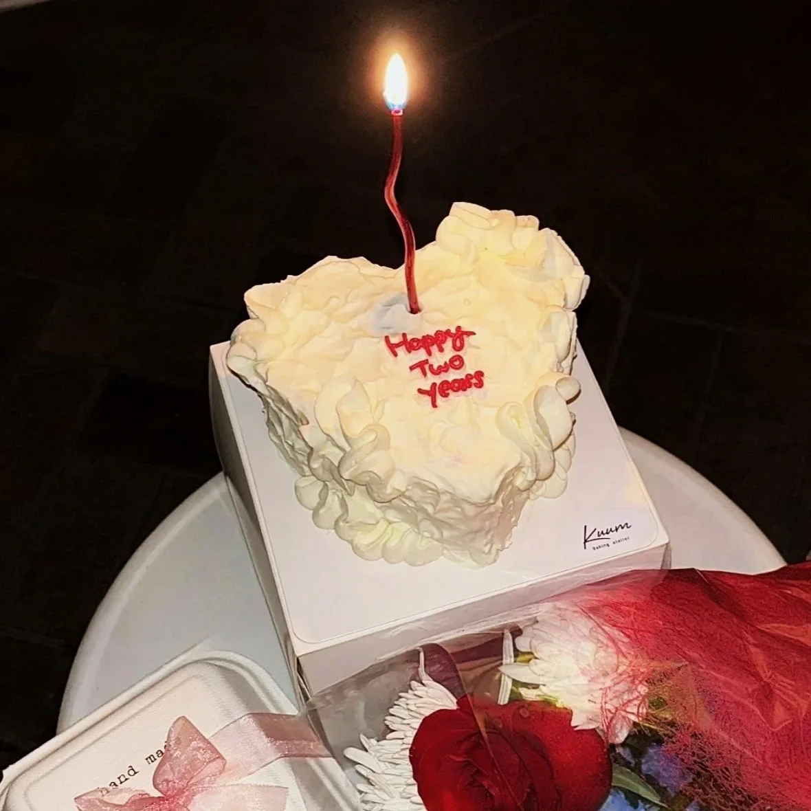 A heart-shaped white birthday cake with a lit candle on top. Red writing on the cake says, "Happy Two Years." The cake is on a white surface, with a red flower and some wrapped items nearby.