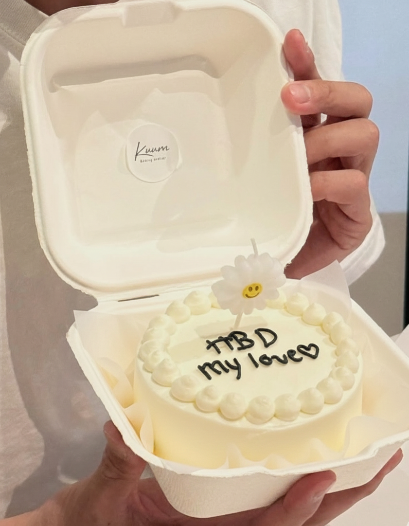 A white frosted cake inside a white takeout box with the message 'TBD my love' written in black icing and a small daisy decoration with a smiley face on top.