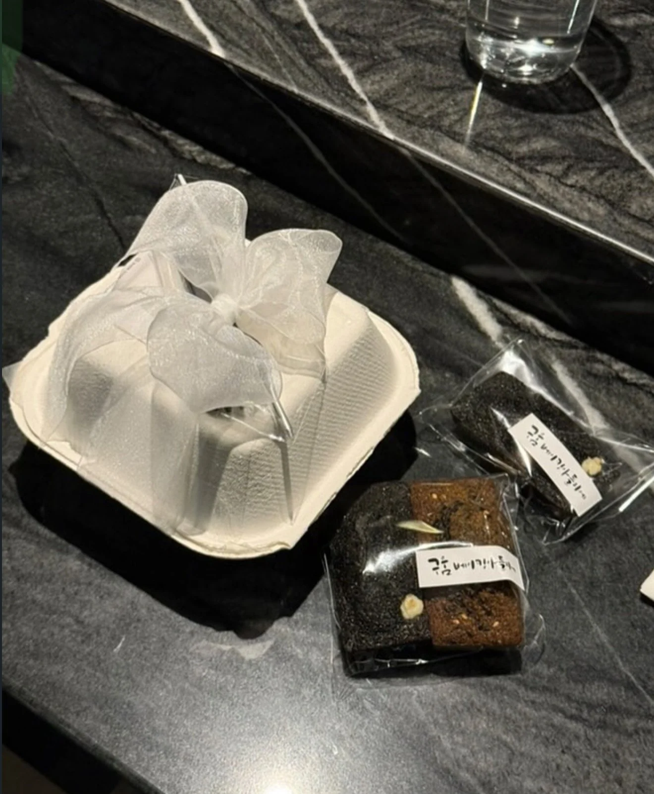 Two decorated sugar cookies on sticks in a white foam tray, and two sealed snack packets with Korean writing, all on a dark countertop with a glass of water