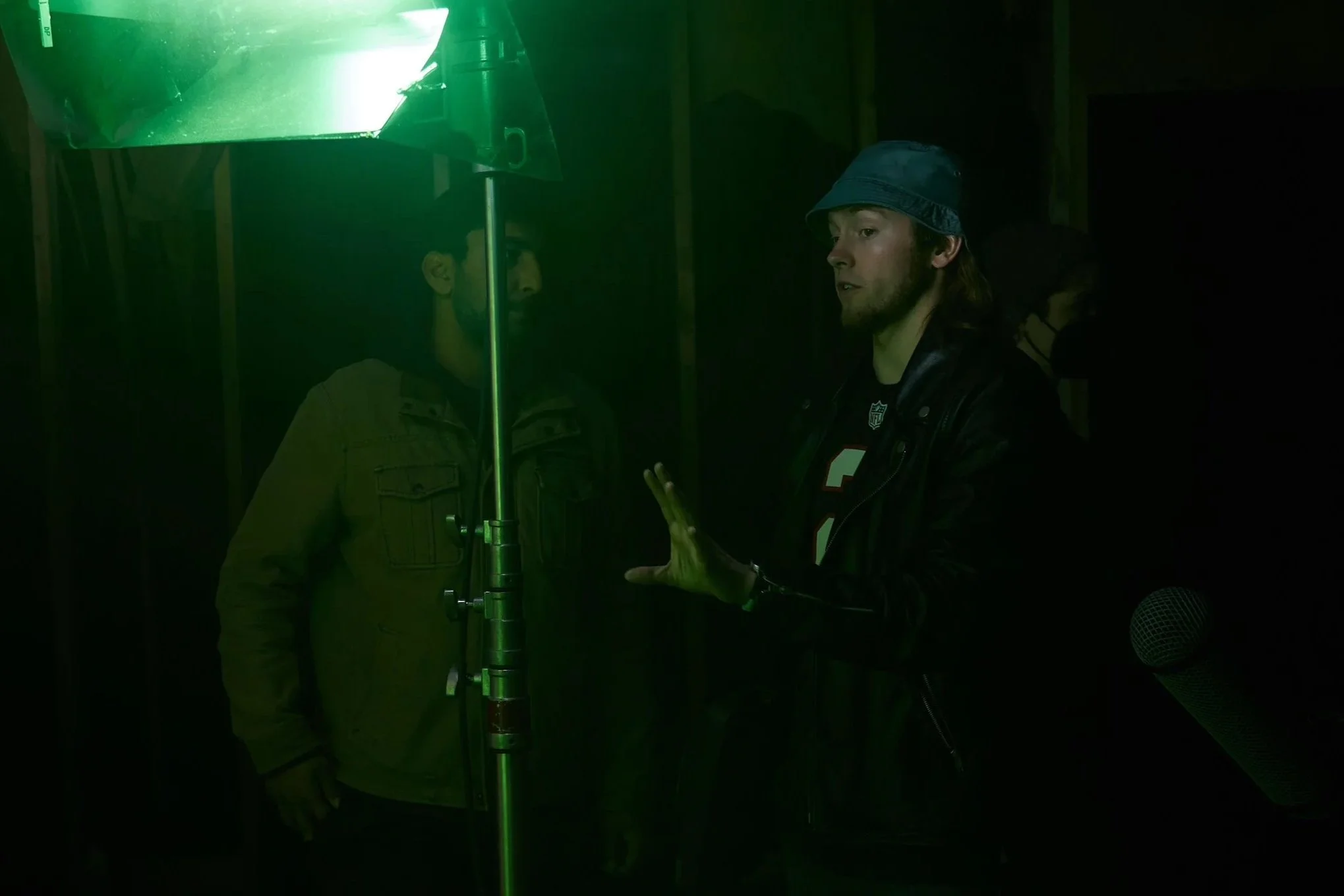 Two men in a dark room with green lighting, one is talking and gesturing with his hand, the other is listening. There is a microphone in the lower right corner.