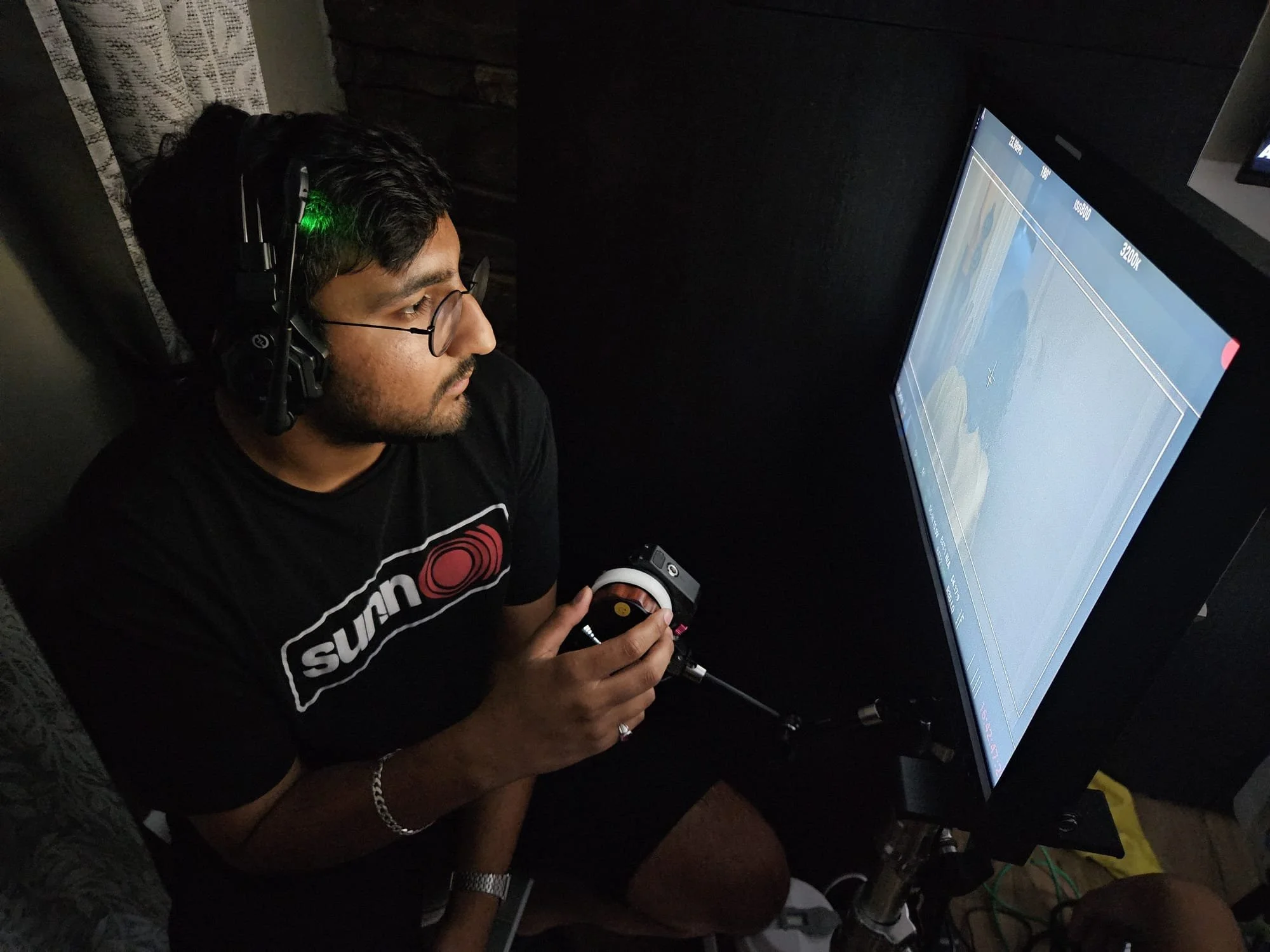 A man with glasses, wearing headphones and a 'Sun' T-shirt, is sitting and using a controller in front of a large computer monitor with a graph on it.