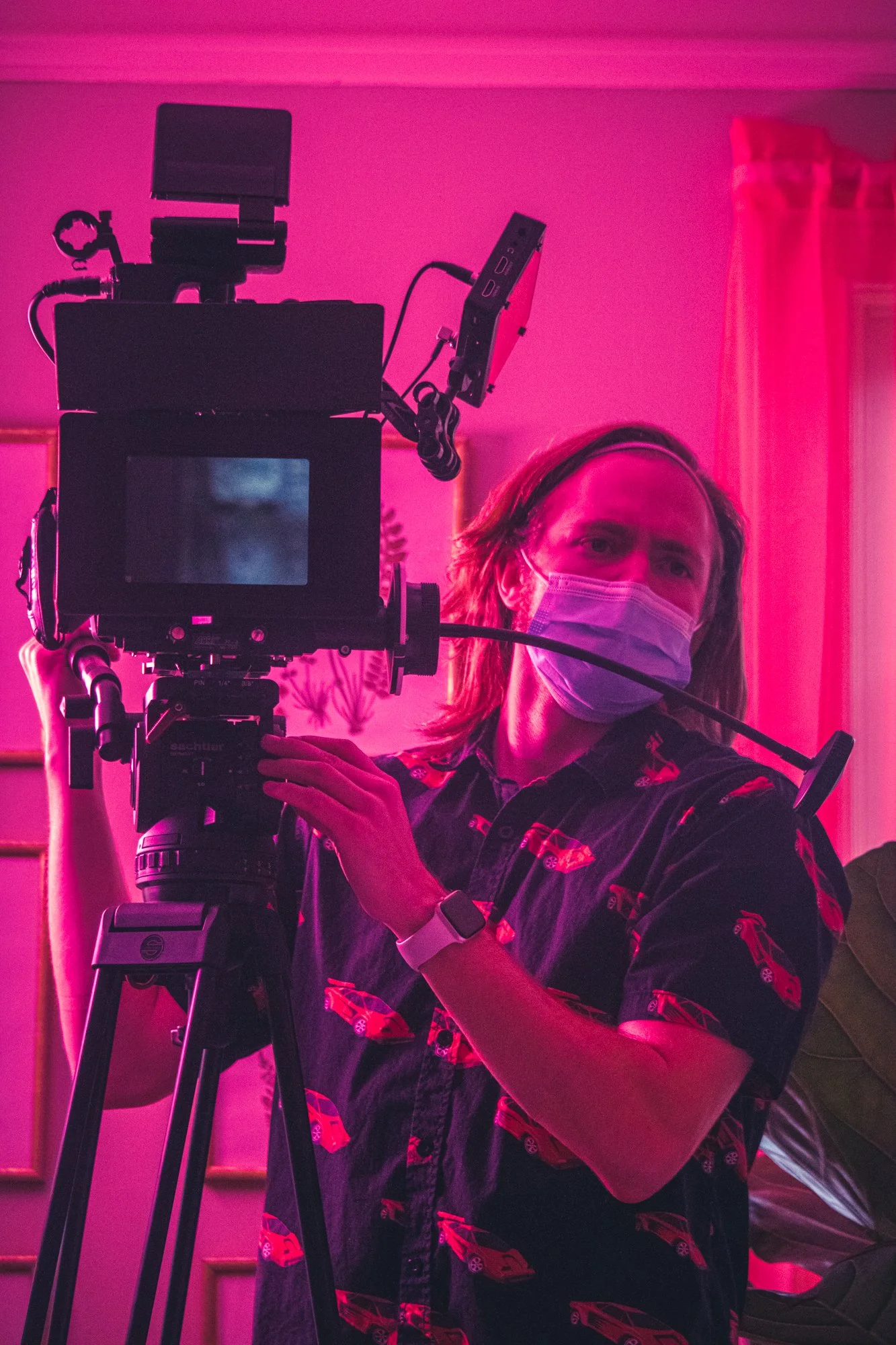 A person wearing a face mask and a black shirt with red cars, operating a professional camera on a tripod indoors with pink lighting.