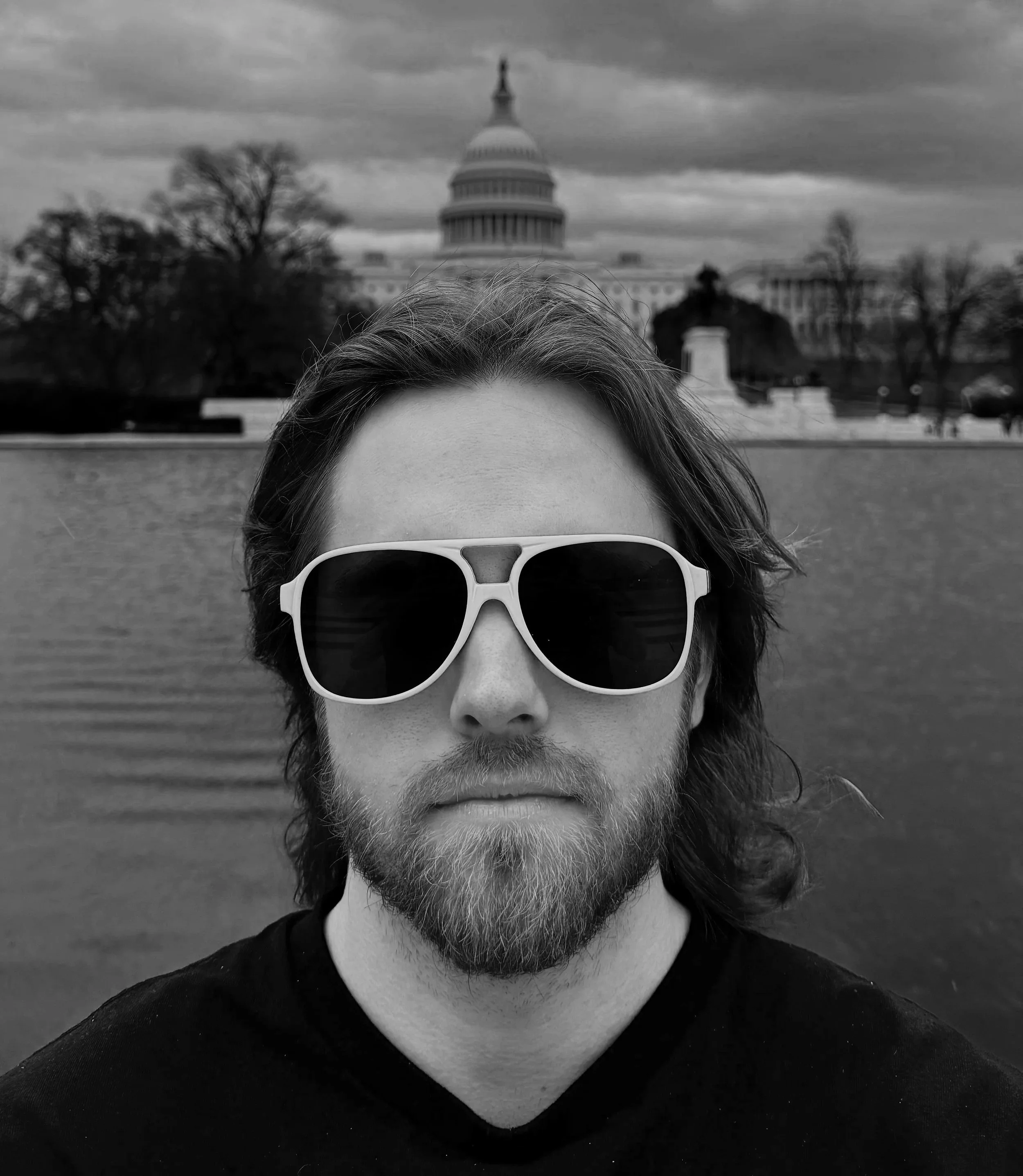 A man with long hair and a beard wearing sunglasses taking a selfie in front of the U.S. Capitol building, with water and trees in the background, in black and white.