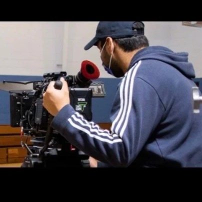 A man in a blue hoodie, black cap, and face mask operating a professional video camera with a red microphone cover.