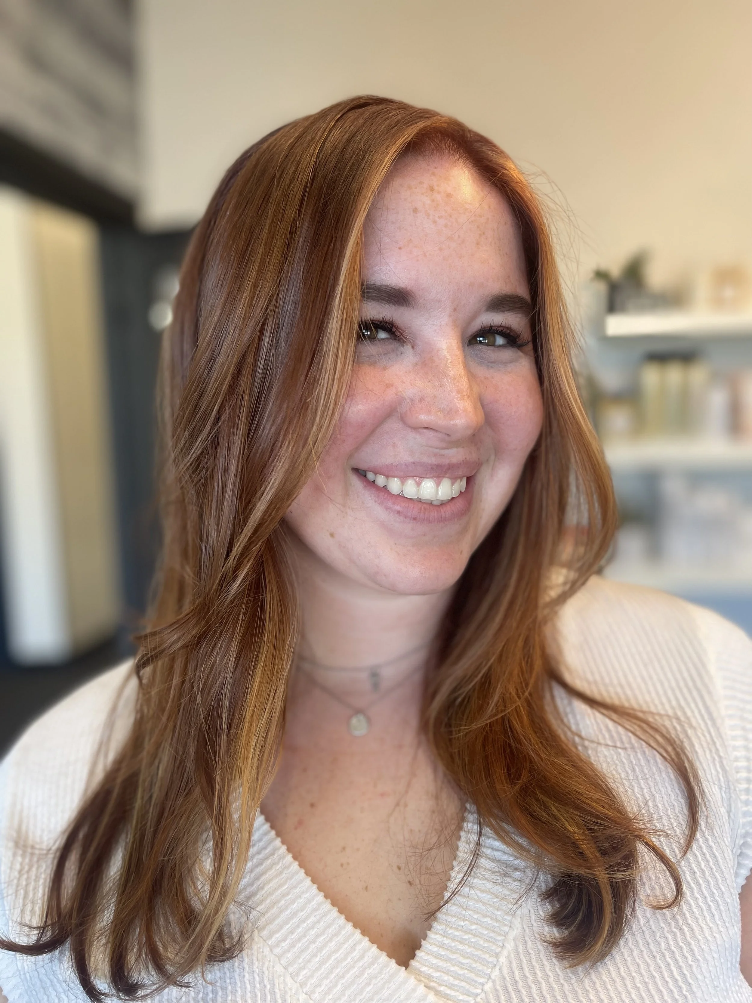 A young woman with long red hair, light skin with freckles, smiling and wearing a white V-neck top and layered necklaces. The background shows a softly blurred interior space with shelves and decor.