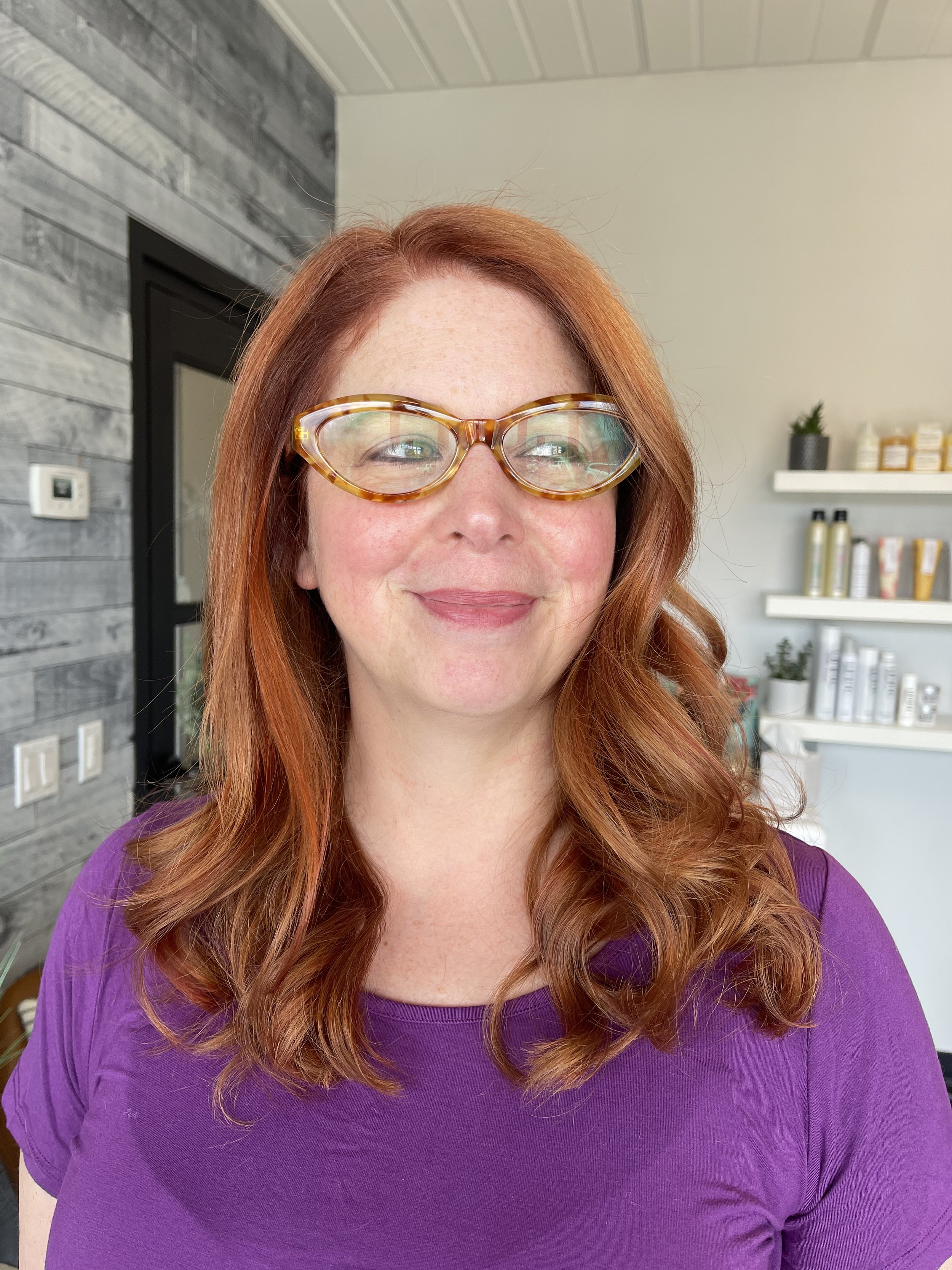 A woman with red hair, wearing glasses with a tortoiseshell pattern, smiles at the camera. She is dressed in a purple shirt, and the background features a modern interior with wooden wall panels, shelves holding skincare or cosmetic products, and som