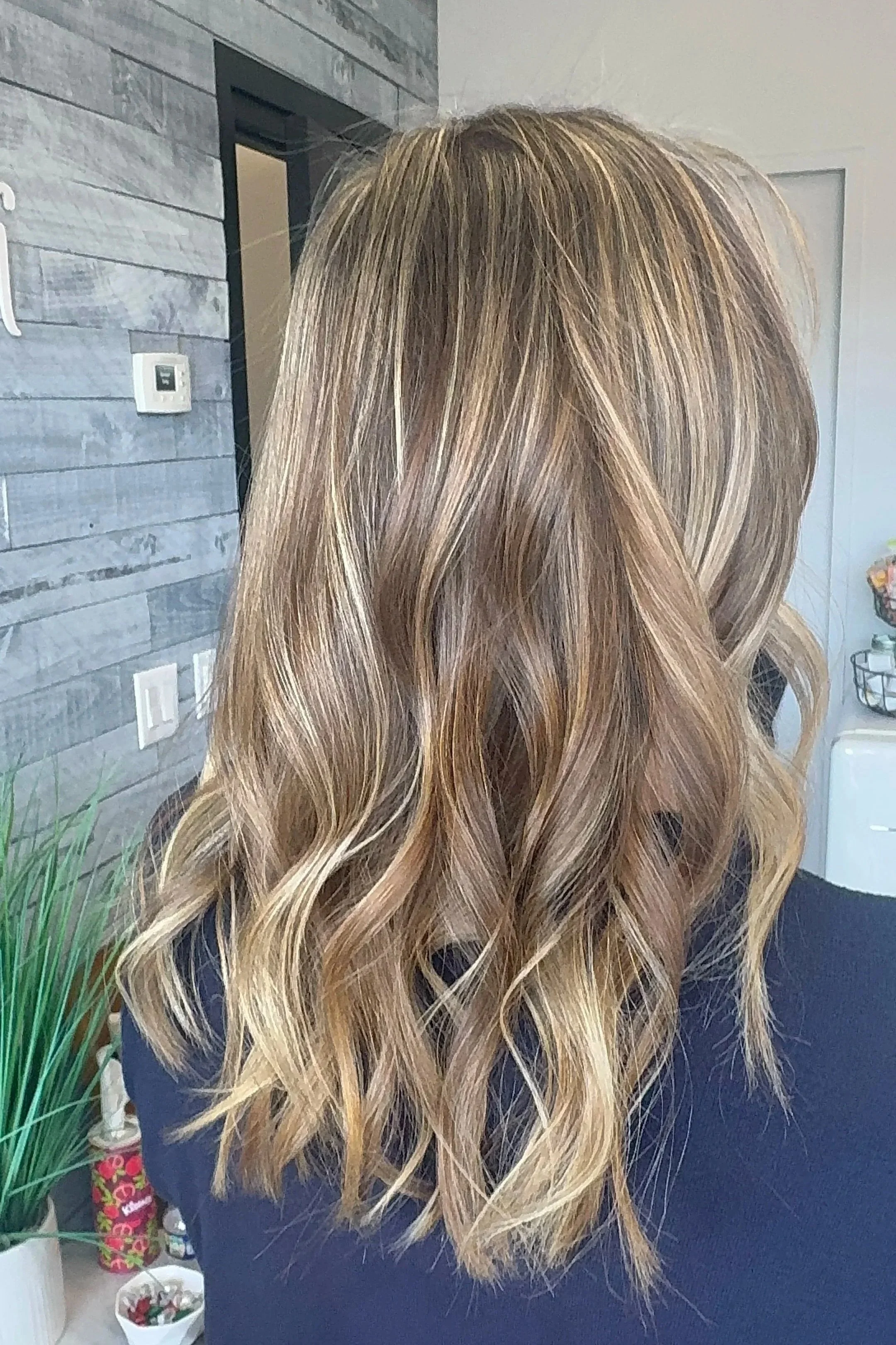 Back of a woman’s head with shoulder-length, wavy, blonde hair with darker roots, indoors.