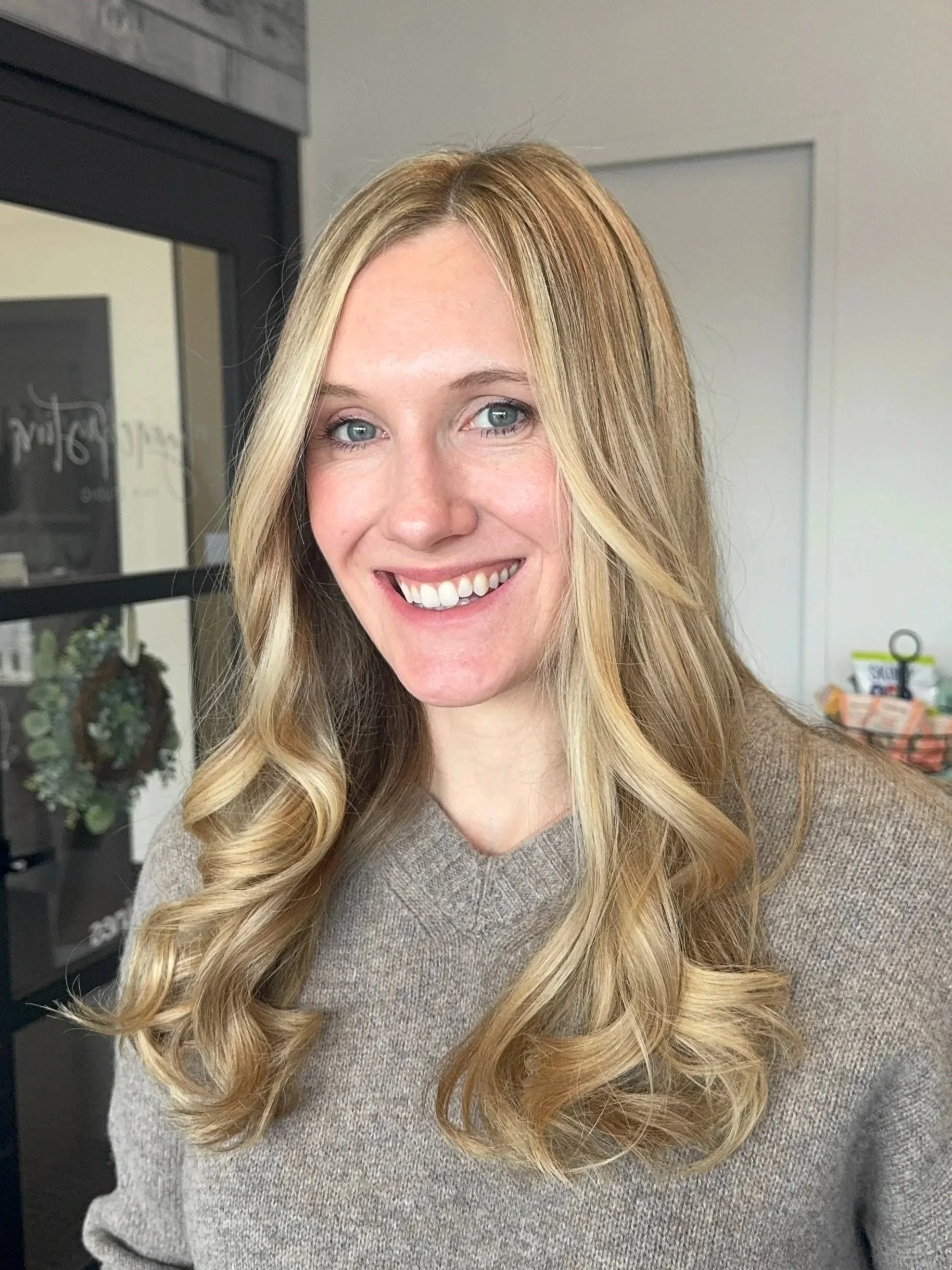 A woman with long blonde hair, blue eyes, and a brown sweater, smiling in a room with a door and some household items in the background.