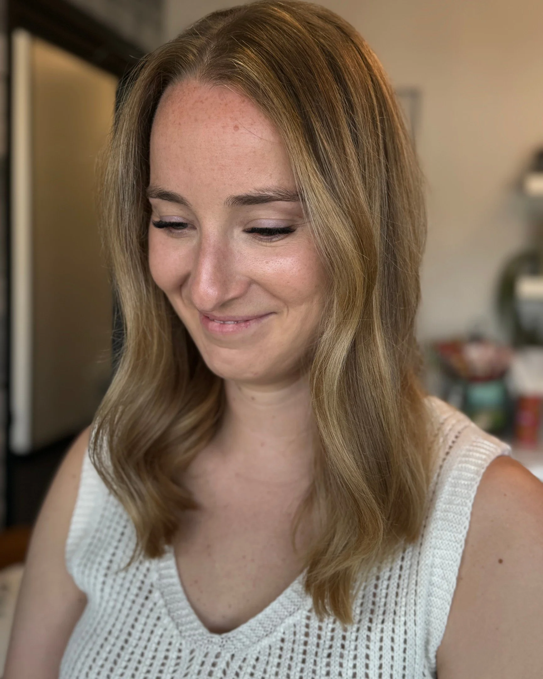 A woman with light skin and shoulder-length wavy blonde hair smiling softly while looking down, wearing a white sleeveless top.