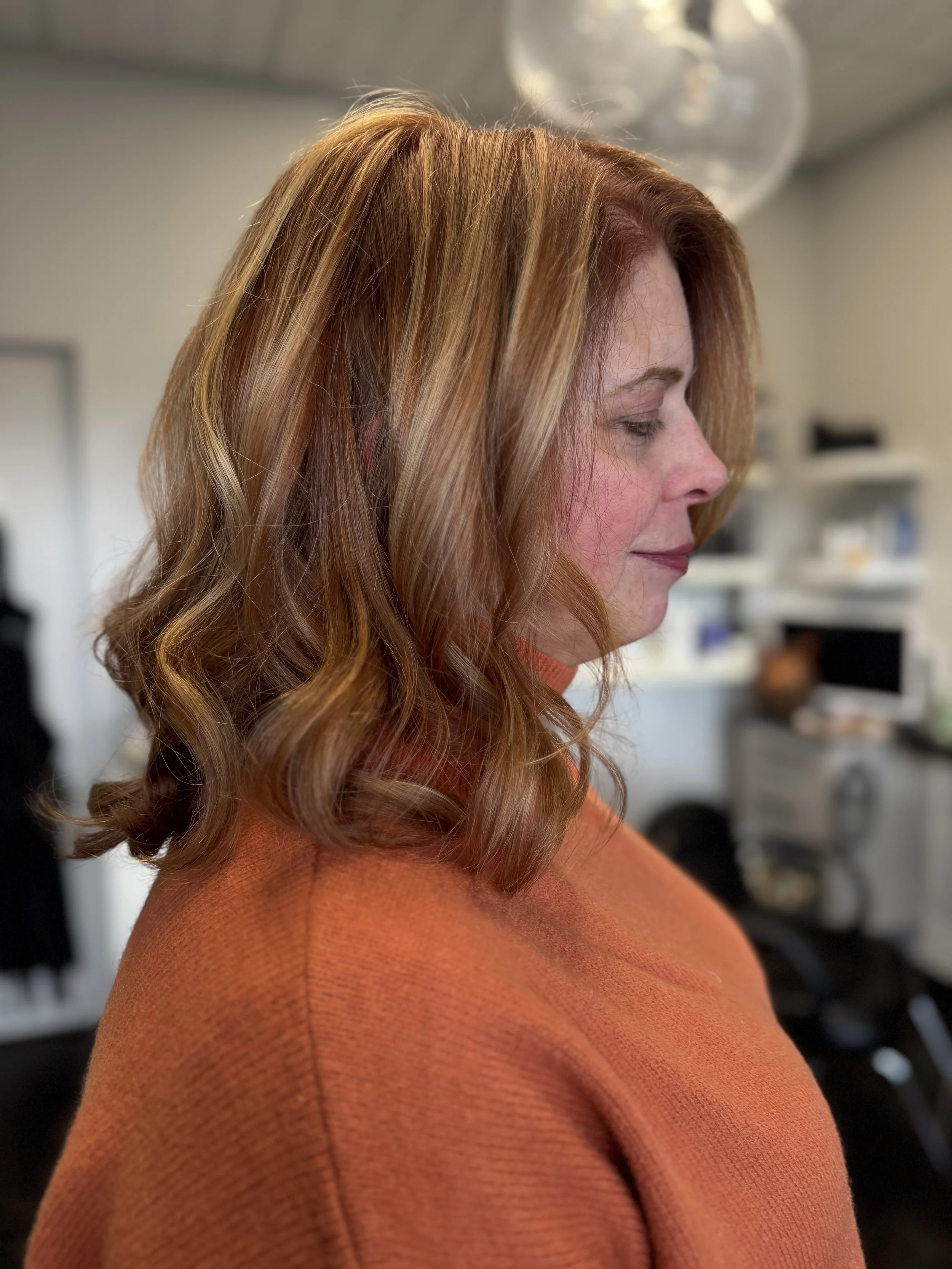 Side profile of a woman with wavy, red hair wearing an orange top, indoors.