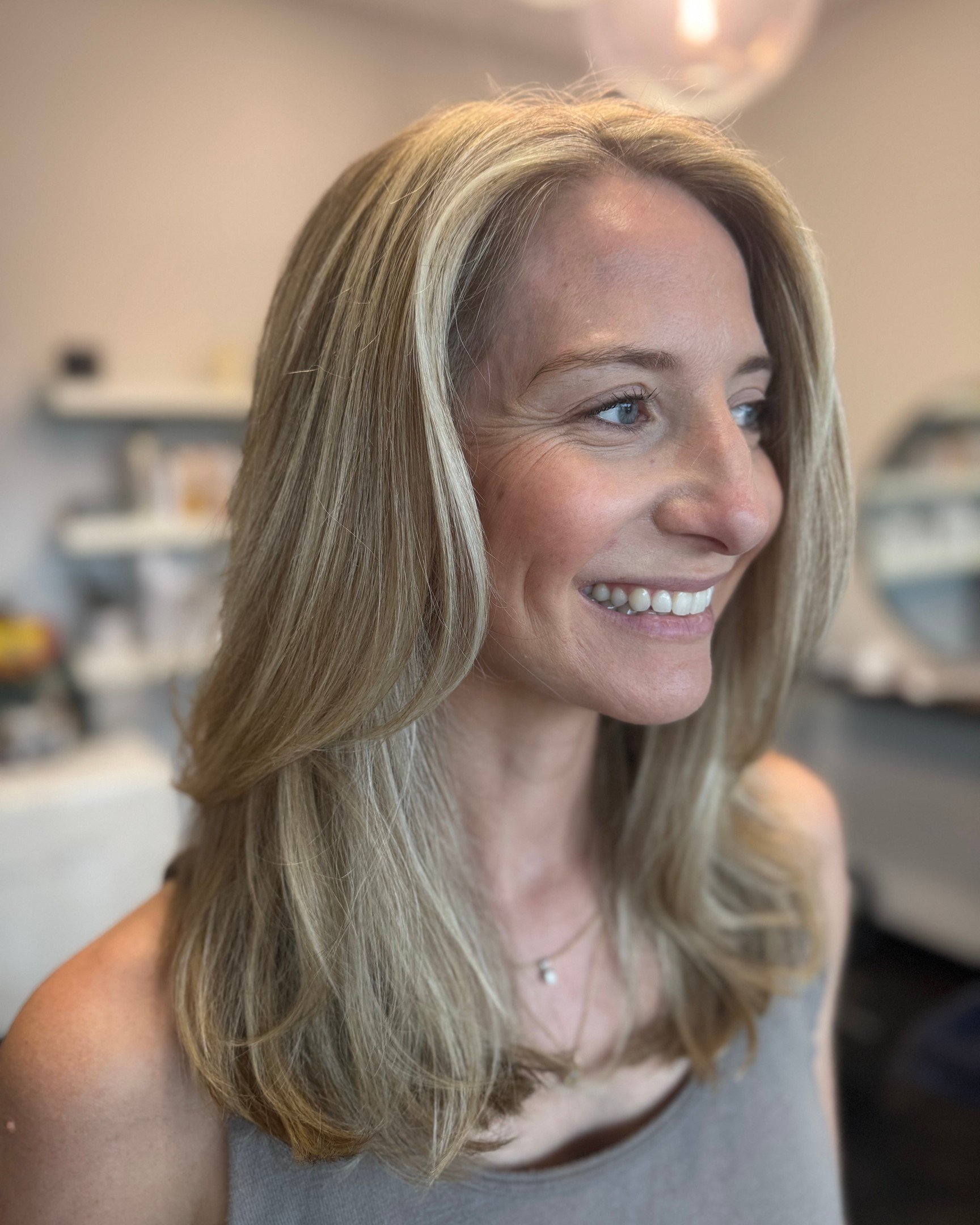 A smiling woman with blonde hair and blue eyes, wearing a gray top and a necklace, in a softly lit indoor setting.