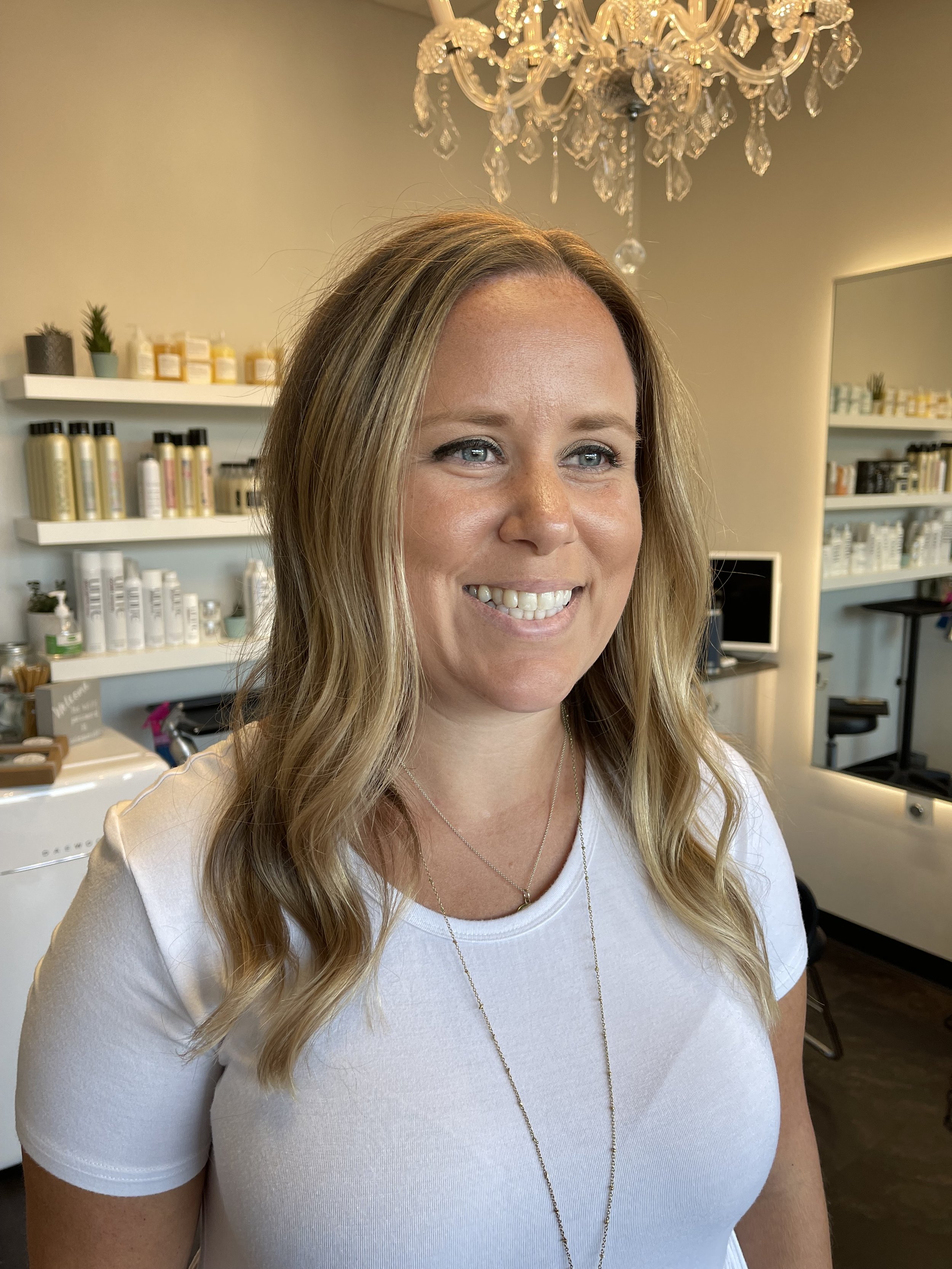 A woman with blonde hair smiling in a room with shelves of bottles and a chandelier overhead.