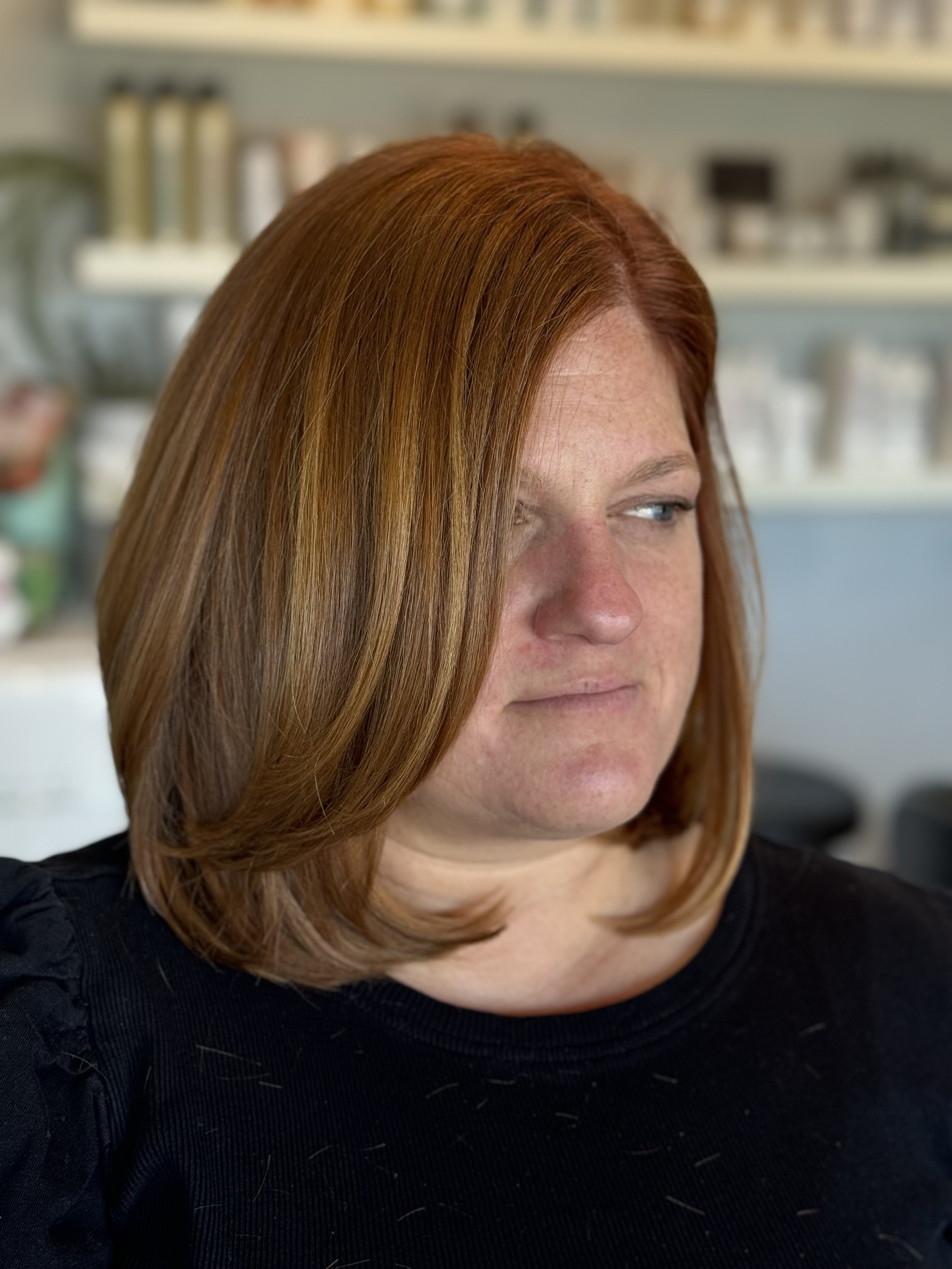 A woman with red hair styled in layers, wearing a black shirt, looking to the side in a salon or spa setting with shelves of hair and beauty products in the background.